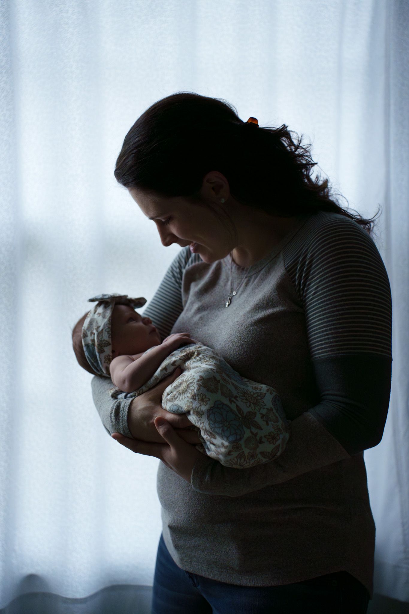 A silhouetted figure in a sweater holds and gazes lovingly at a newborn baby near a bright window.