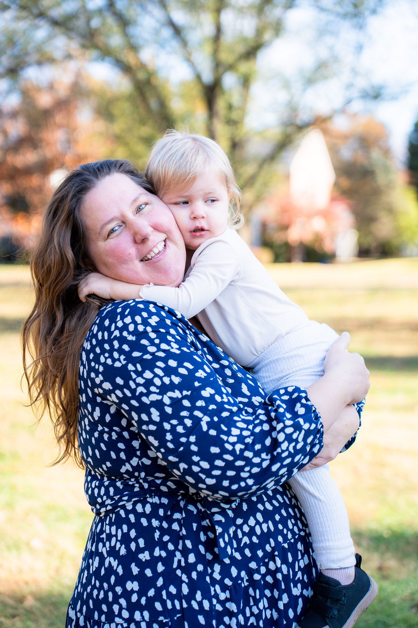 A tender outdoor moment captured in warm sunlight with a navy polka dot dress against a natural autumn backdrop.