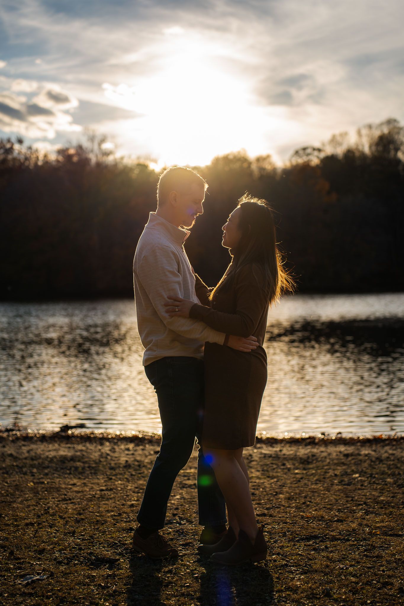 A couple shares an intimate silhouetted moment by a lake at sunset with trees in the background.