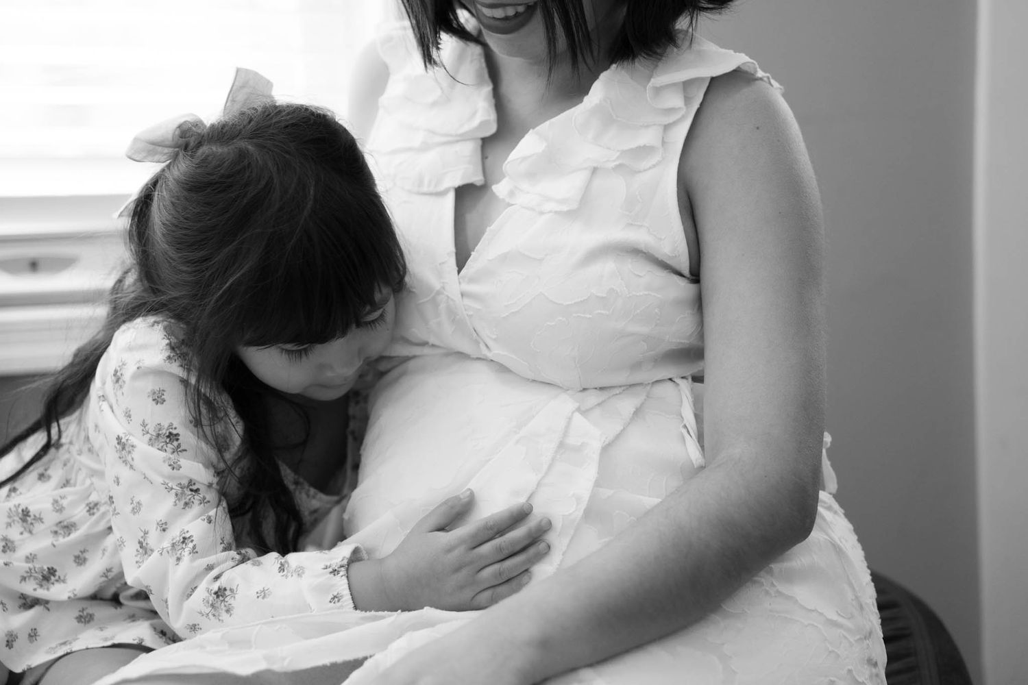 Black and white photo of a young girl kissing a pregnant woman's belly in a tender maternity moment.