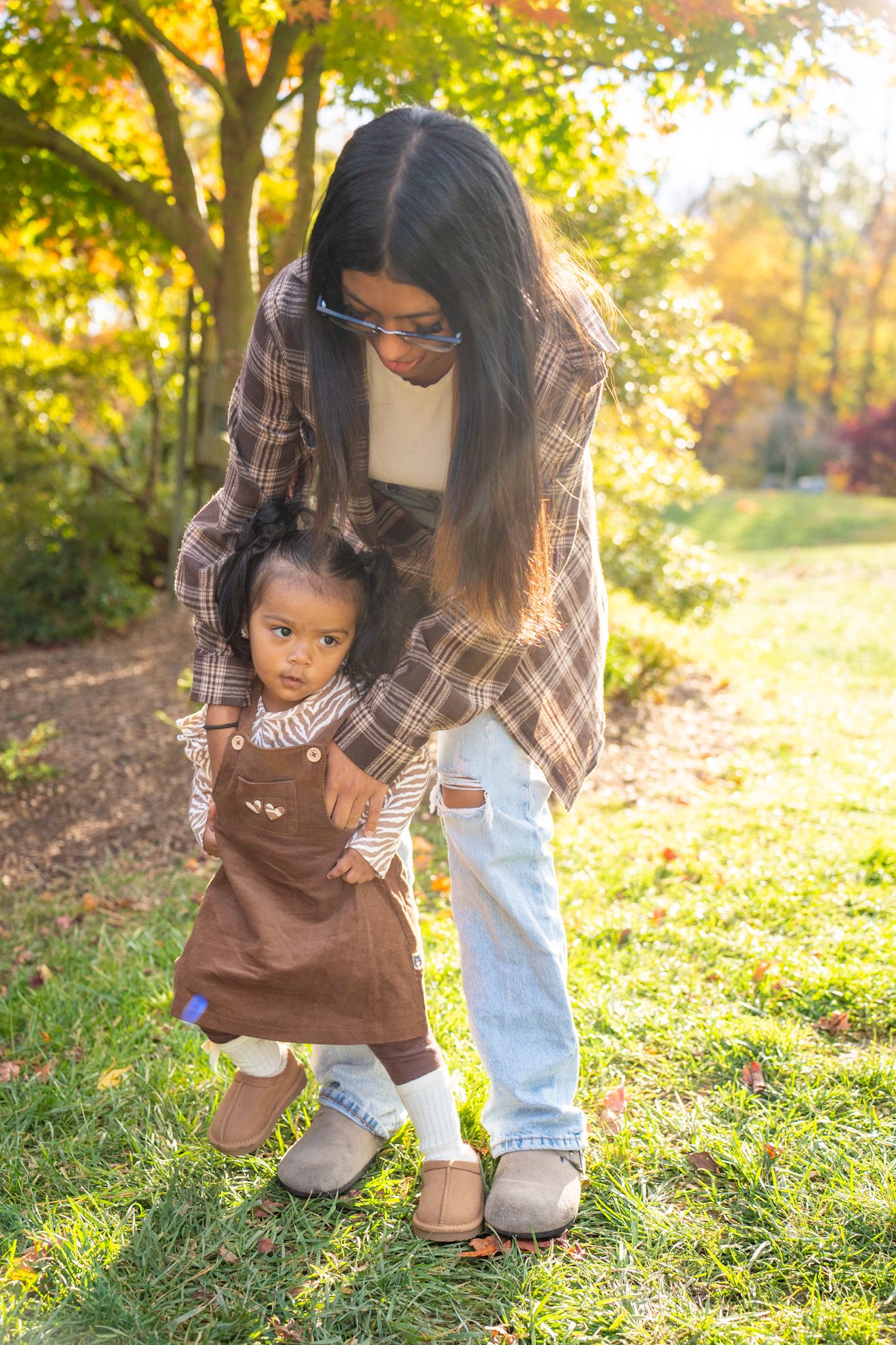 Parent and child explore autumn leaves together on a sunny day in a park setting with golden light filtering through trees.