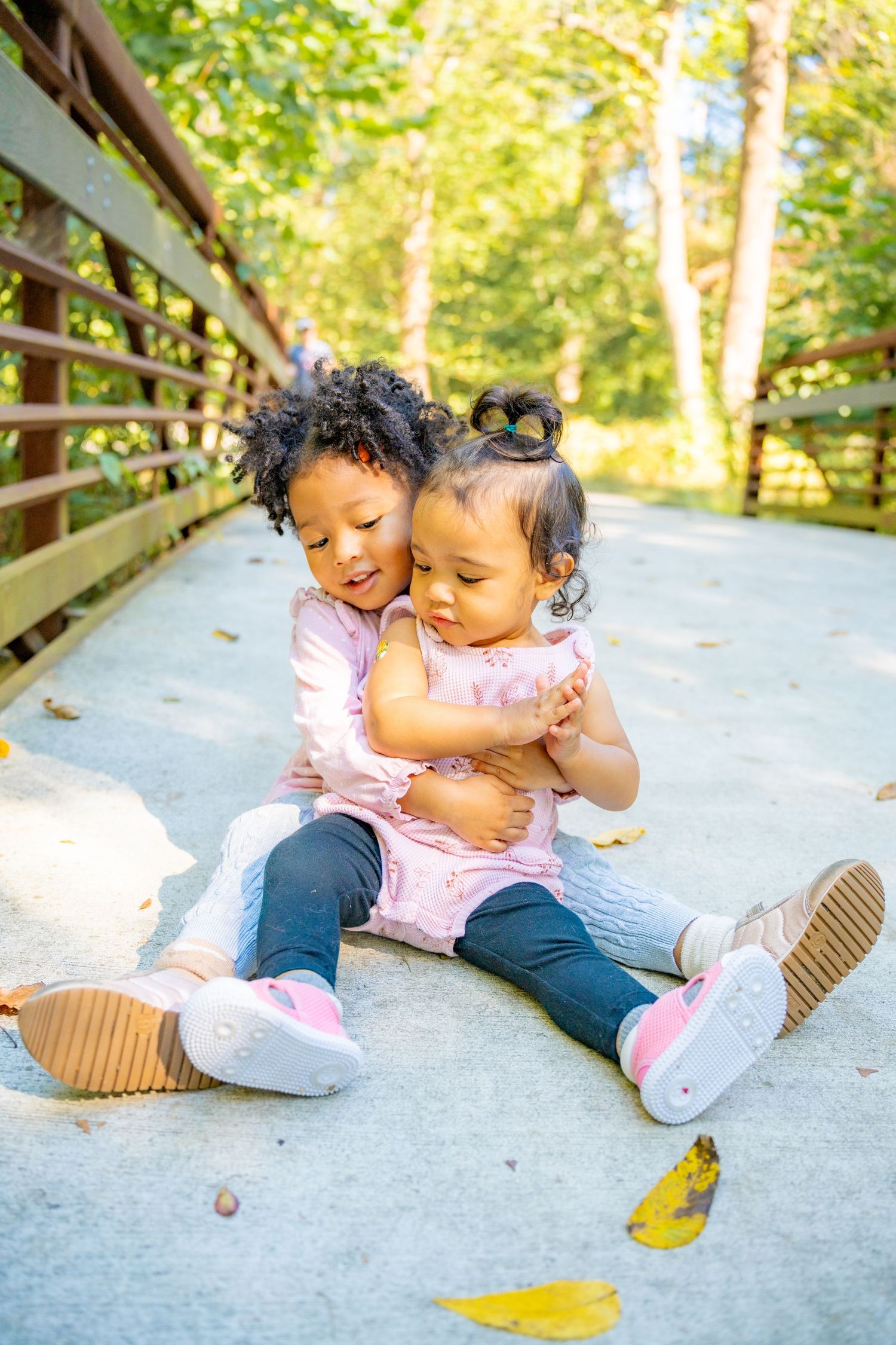 Two toddler siblings sit together laughing and hugging on a wooden bridge path surrounded by green trees.