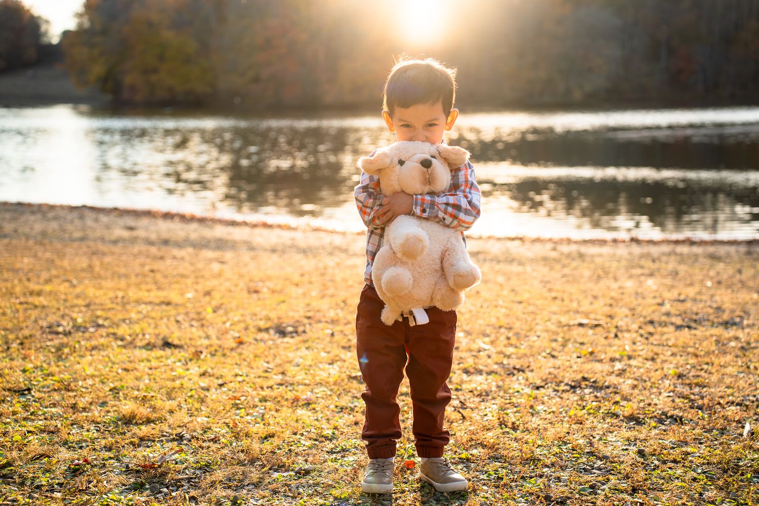 Young boy holding a stuffed teddy bear outdoors by a lake during golden hour in autumn.