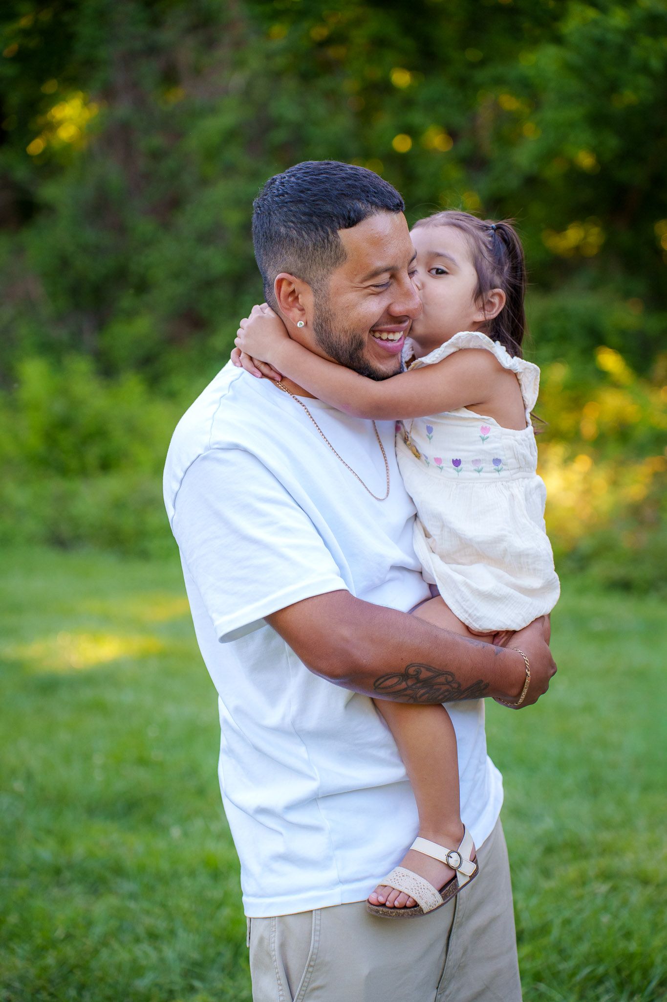 A father holds his young daughter outdoors as she kisses his cheek, both wearing white in a lush green park.