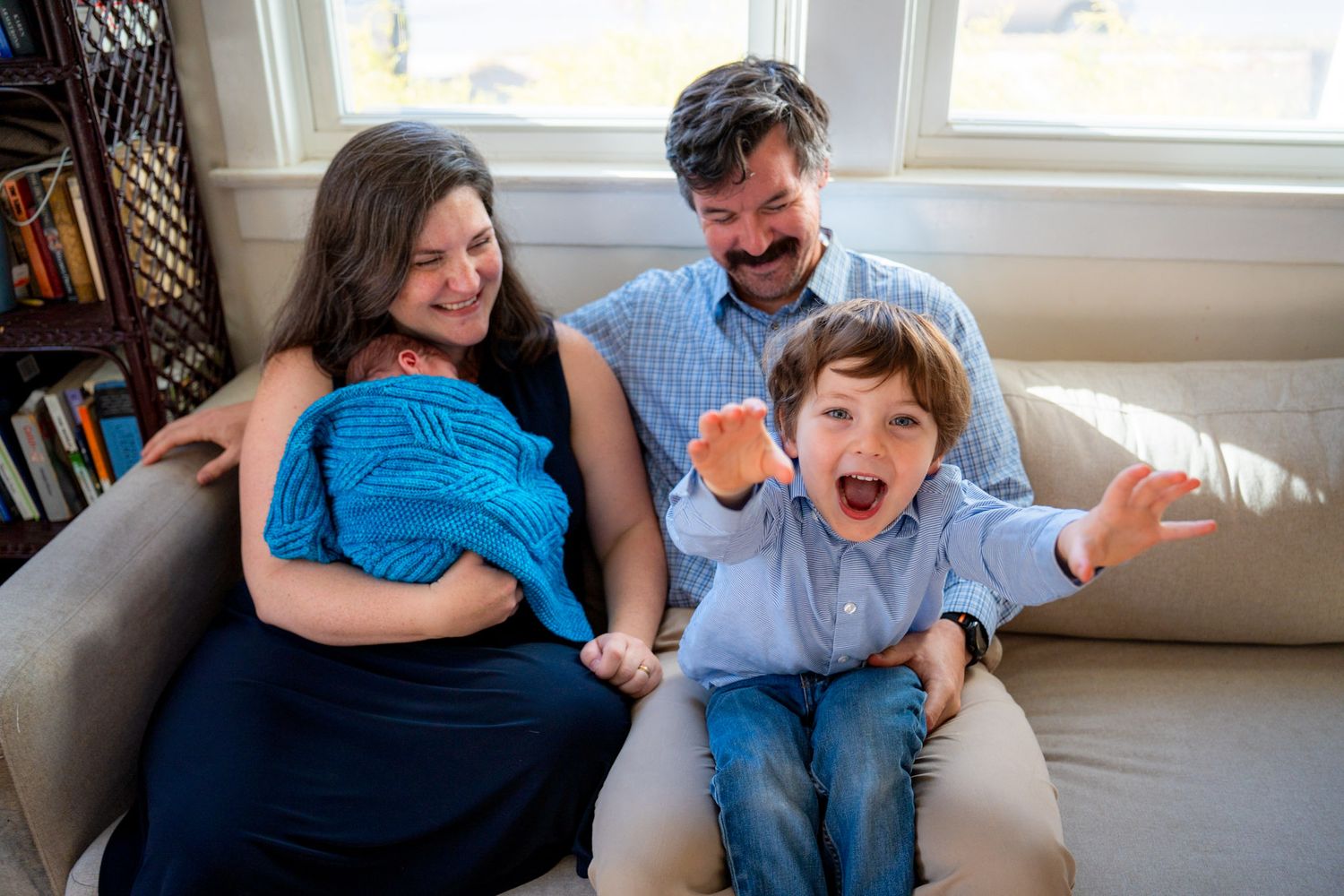 A family relaxes and laughs together while sitting on a gray couch in a bright living room. Silver Spring Lifestyle Newborn Photographer