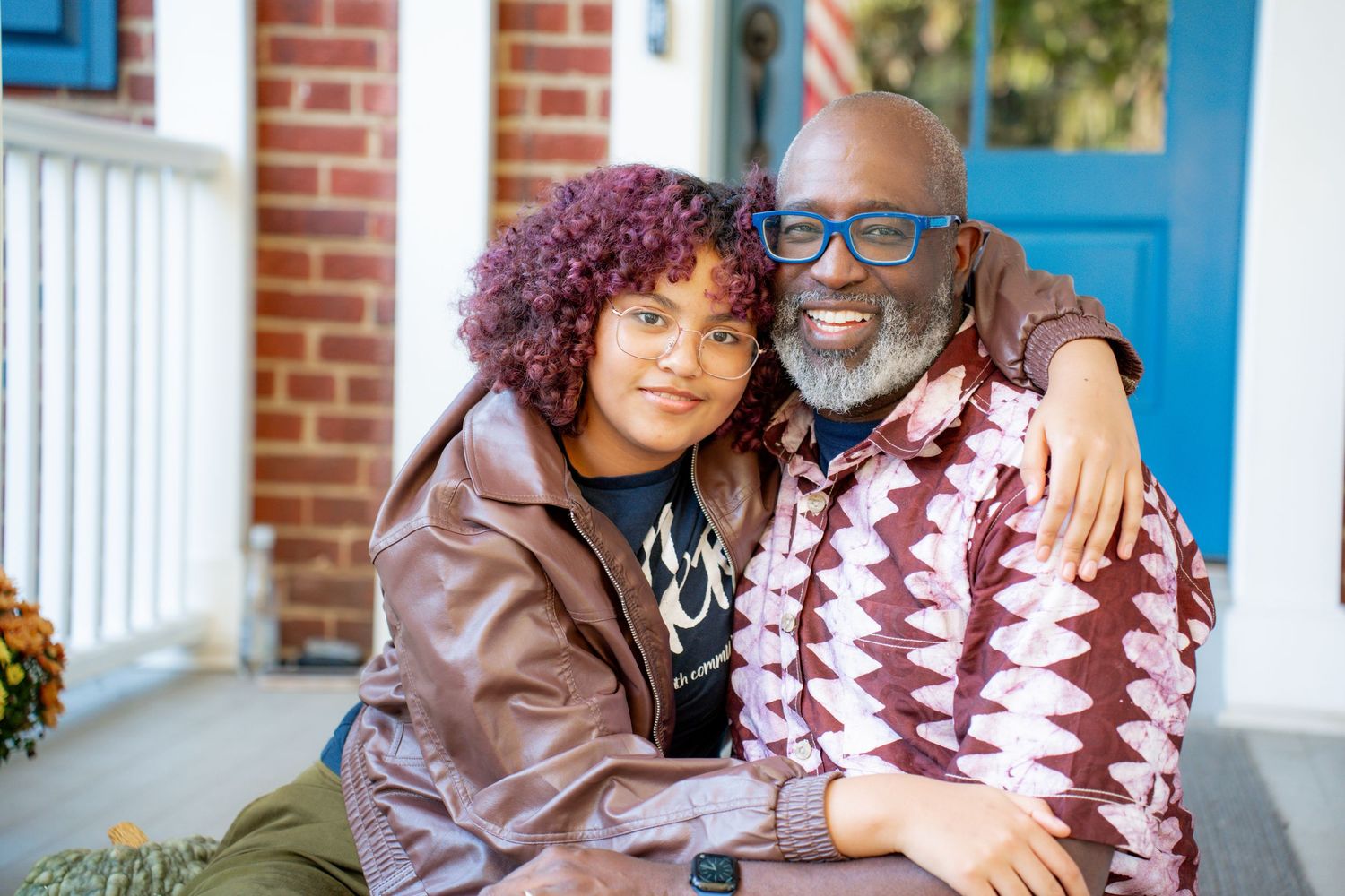 A couple sits together on a porch, wearing cozy fall clothing including a brown leather jacket and patterned sweater.
