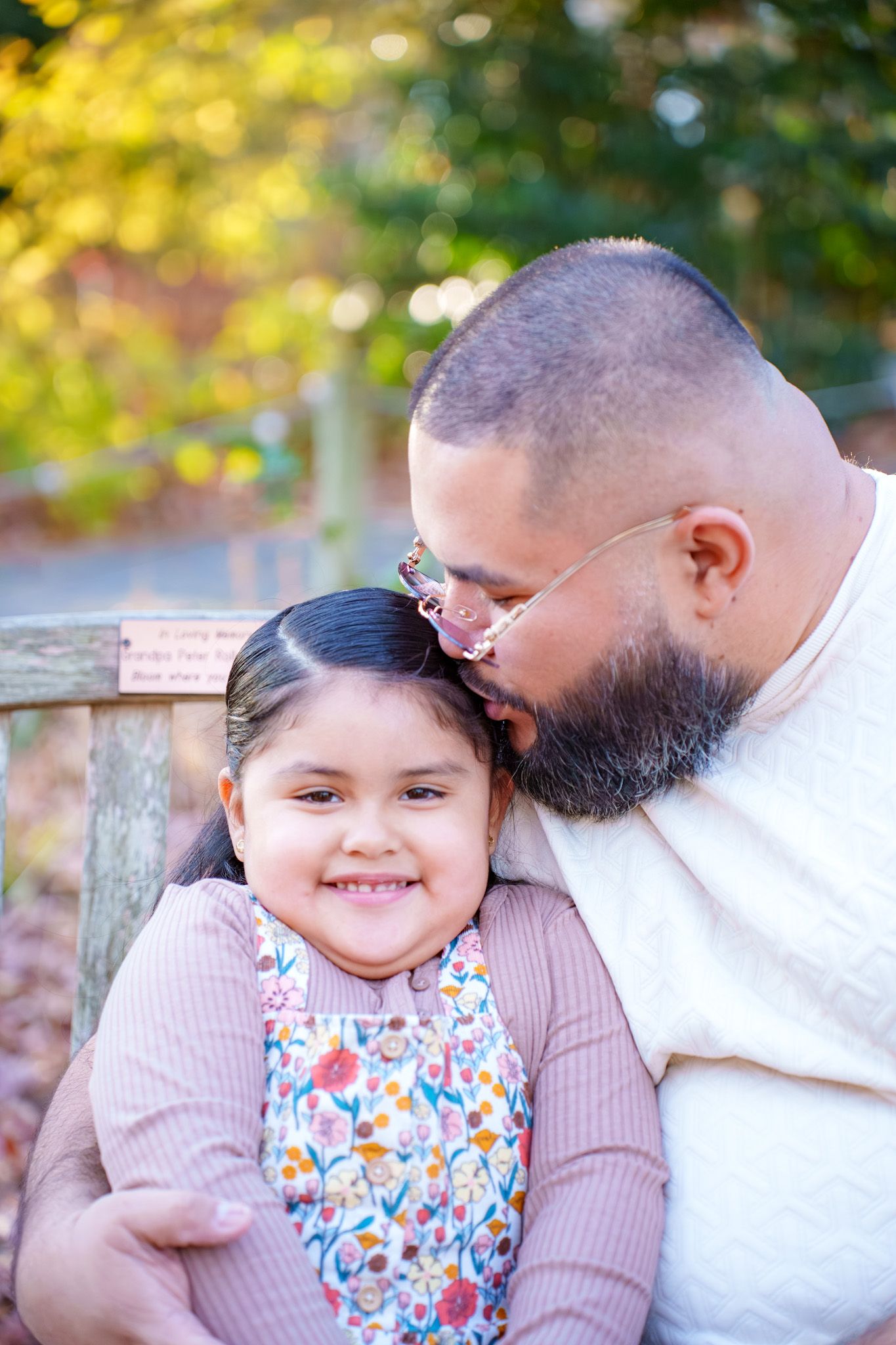 A bearded father lovingly kisses his smiling young daughter on the head while sitting on a bench outdoors.