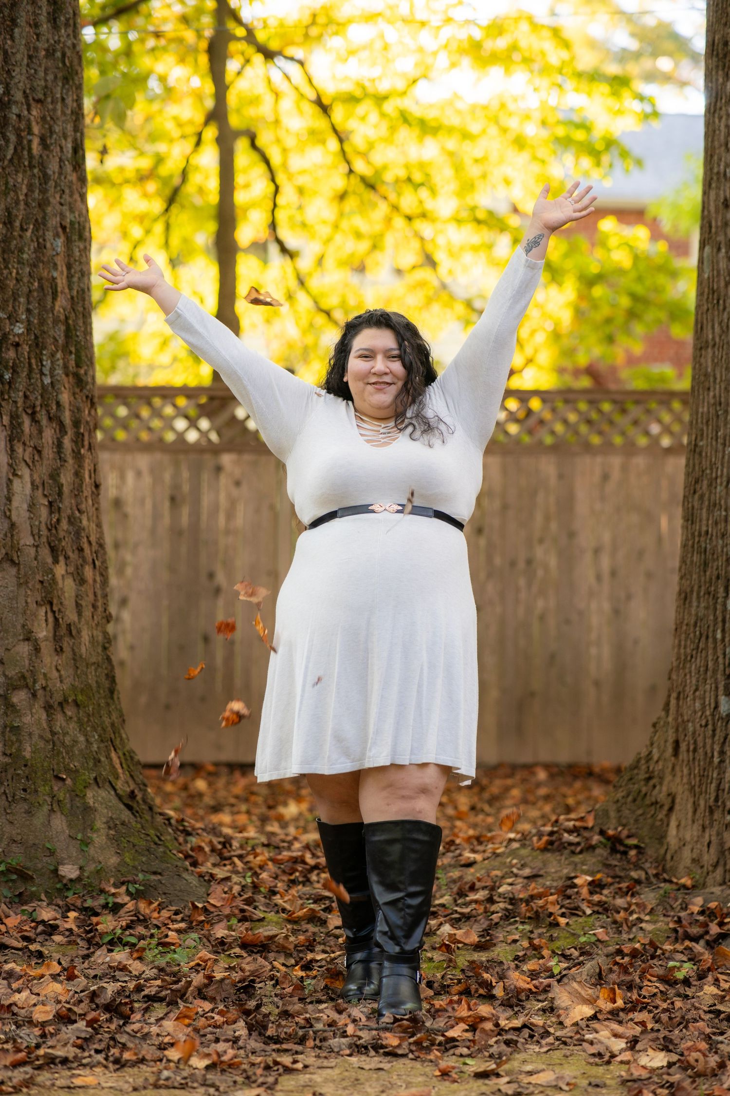 A person in white clothing raises their arms joyfully among fallen leaves and trees during golden hour.