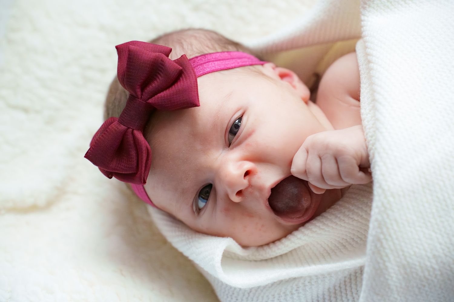 Baby wearing a burgundy bow headband lies on white blanket in four similar poses.
