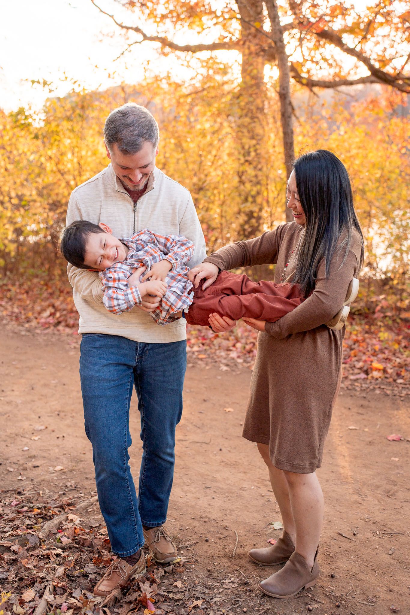 A family shares a playful moment together on a dirt path surrounded by fall colors and fallen leaves.
