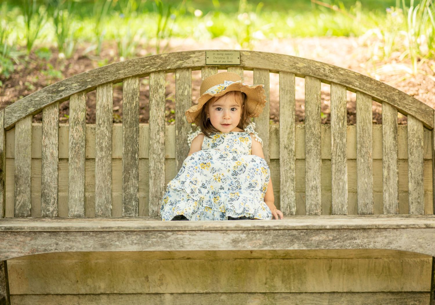 Young girl in floral dress and straw hat sitting on a wooden garden bench surrounded by greenery.