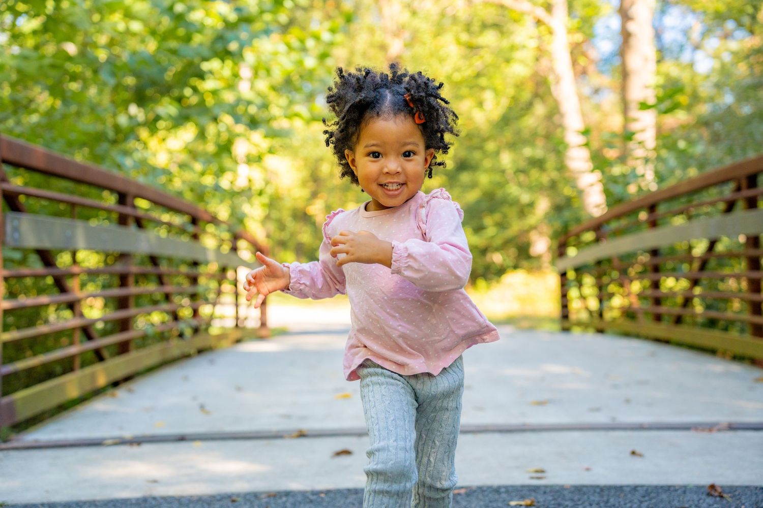 Child in a pink sweater plays and poses on a wooden bridge walkway surrounded by green trees on a sunny day.