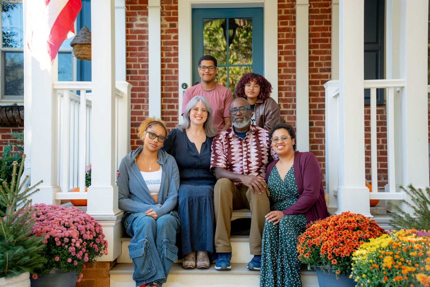 A group of people sit together on a front porch decorated with colorful autumn mums and pumpkins.