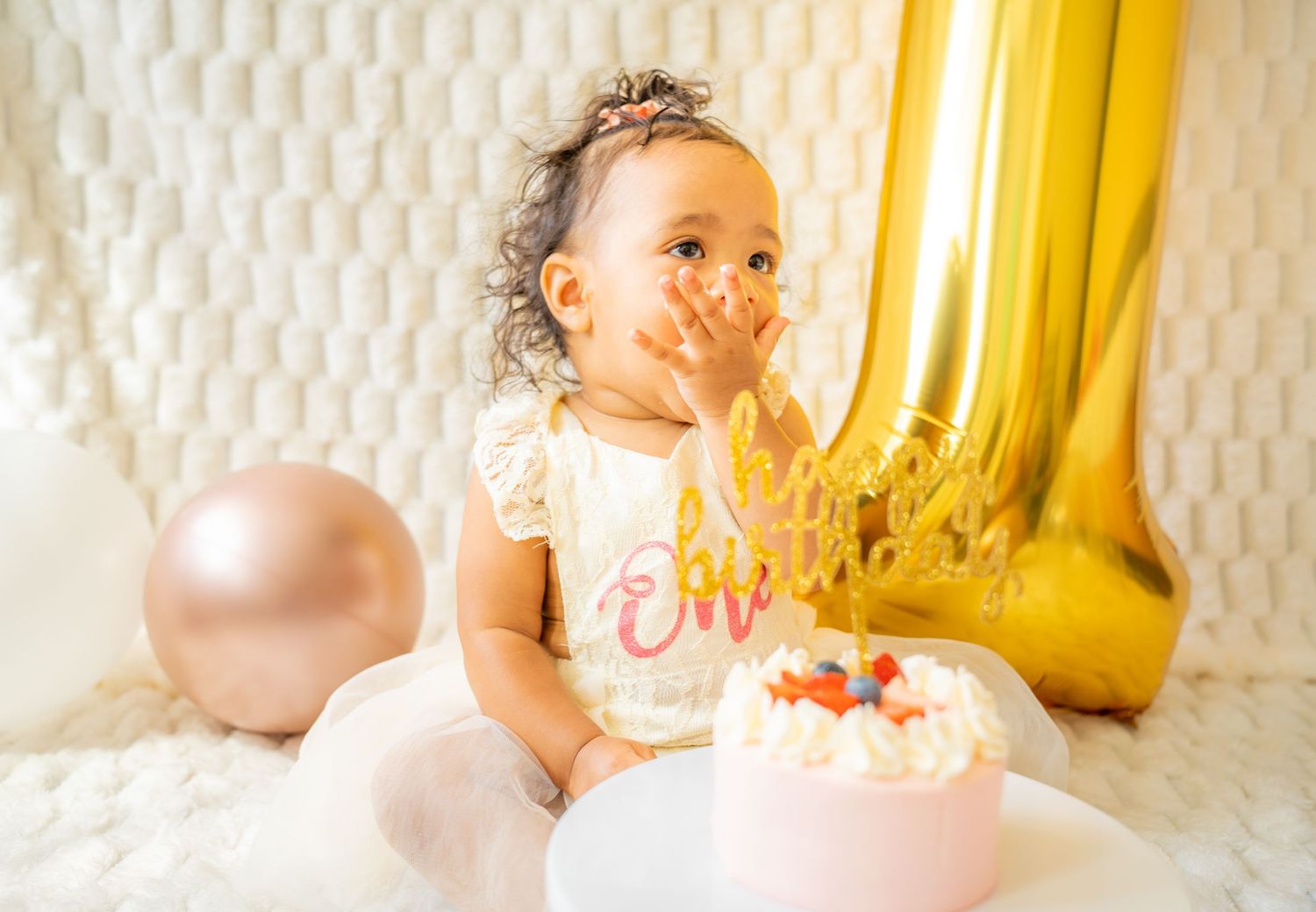 Baby in white dress sitting with birthday cake and gold number one balloon decoration.