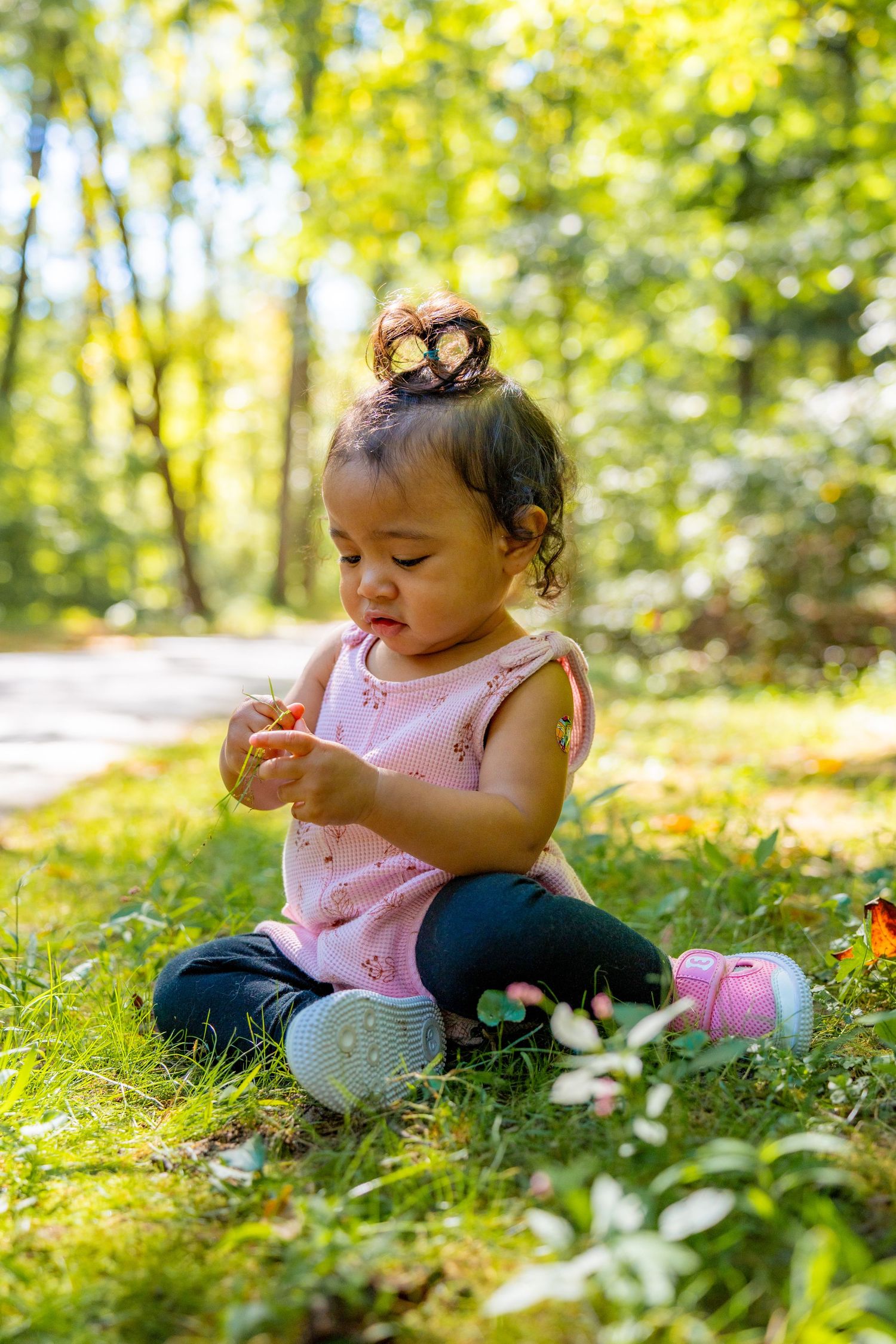 A toddler in a pink dress sits on grass in a sunlit forest while playing with a small toy.