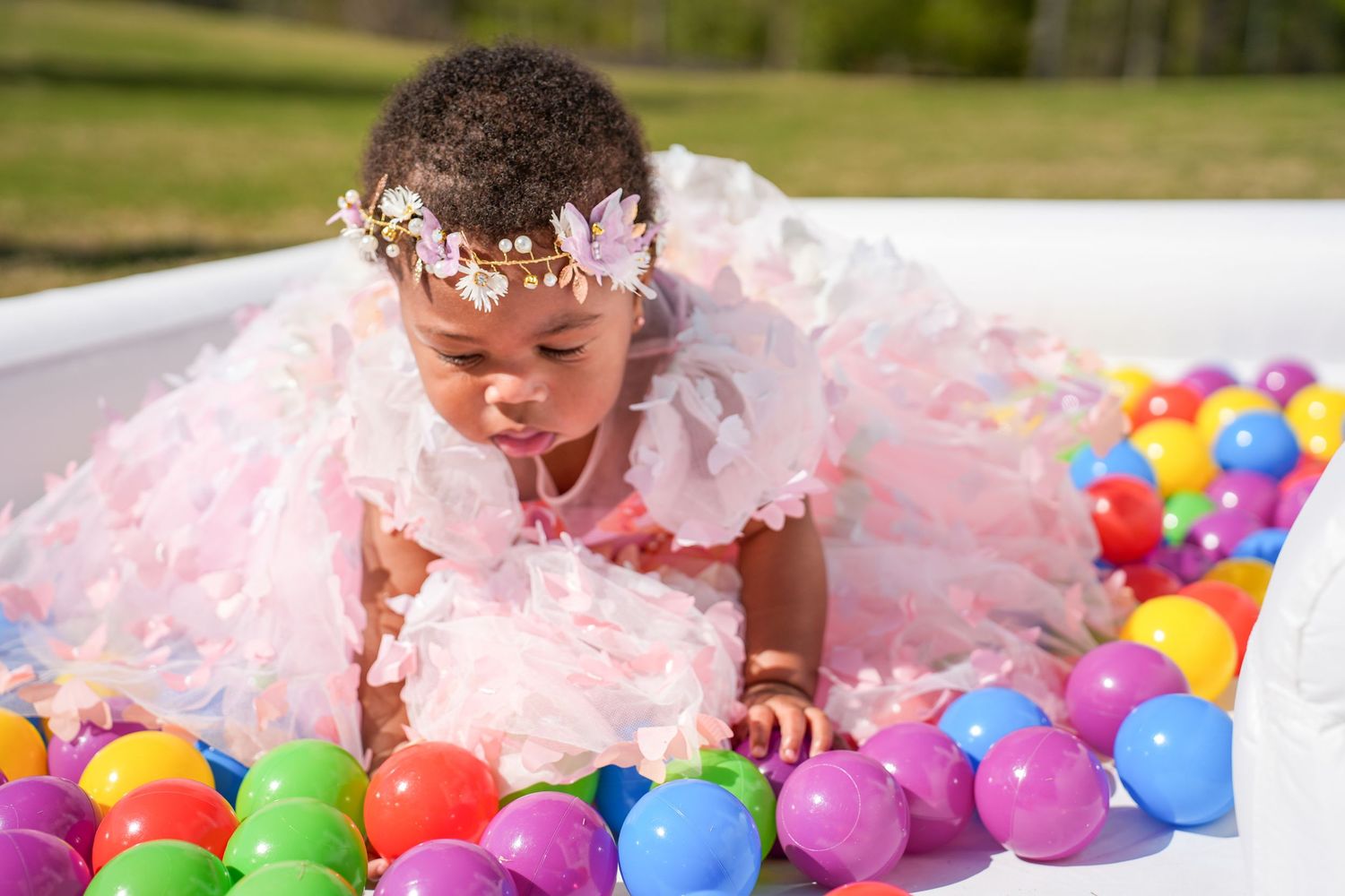 Baby in pink tulle dress crawls through colorful ball pit during first birthday cake smash photo session.