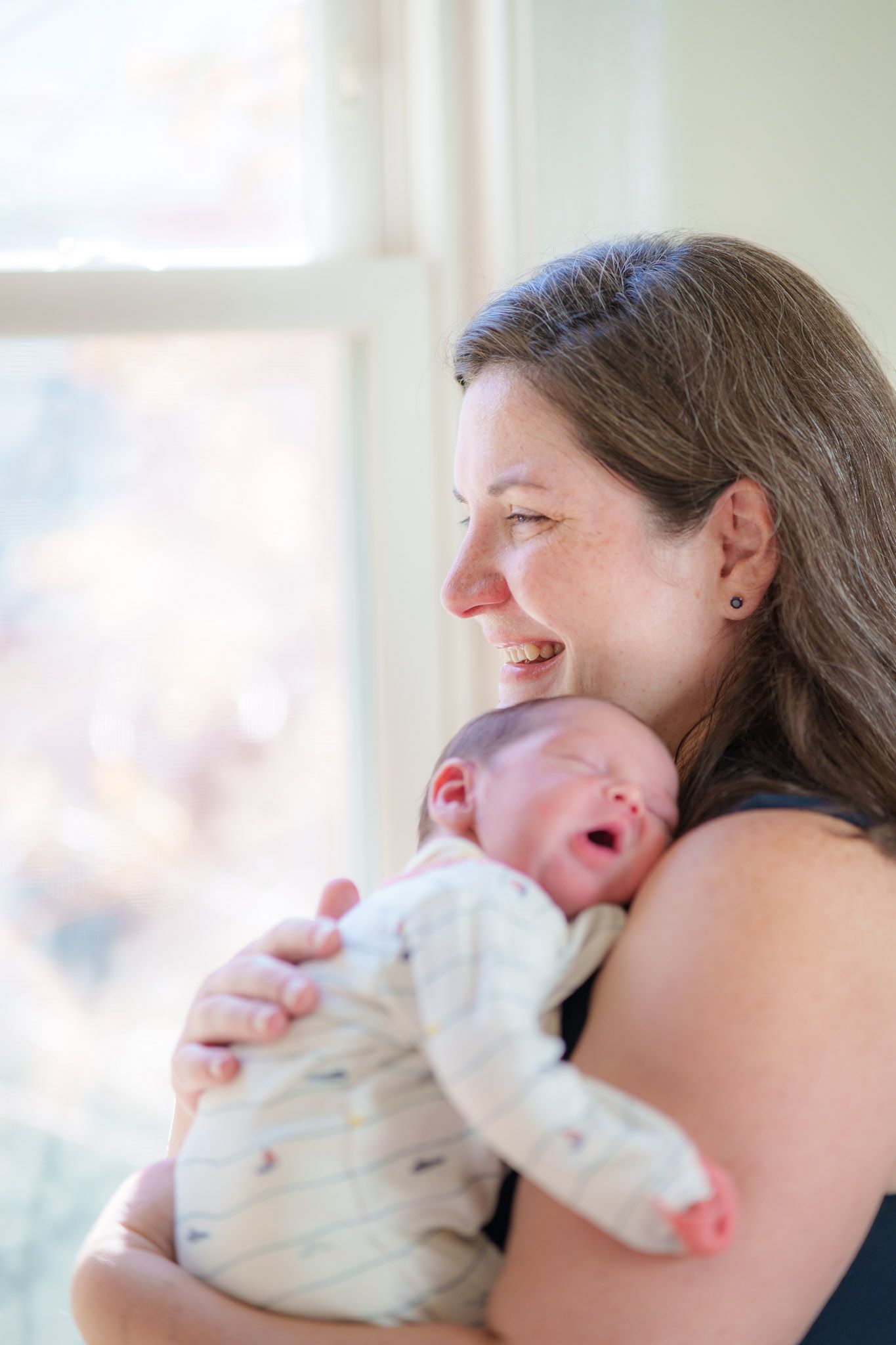 A tender moment between parent and newborn baby by a sunlit window in a peaceful indoor setting.