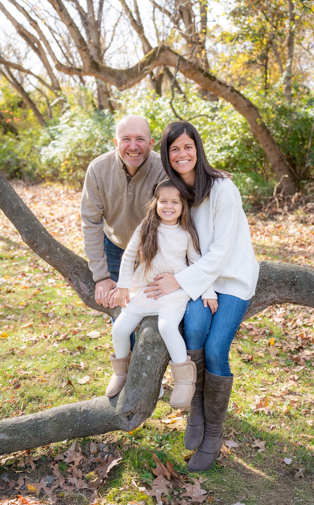 A family of three sits together on a large tree branch during a fall outdoor photo session.