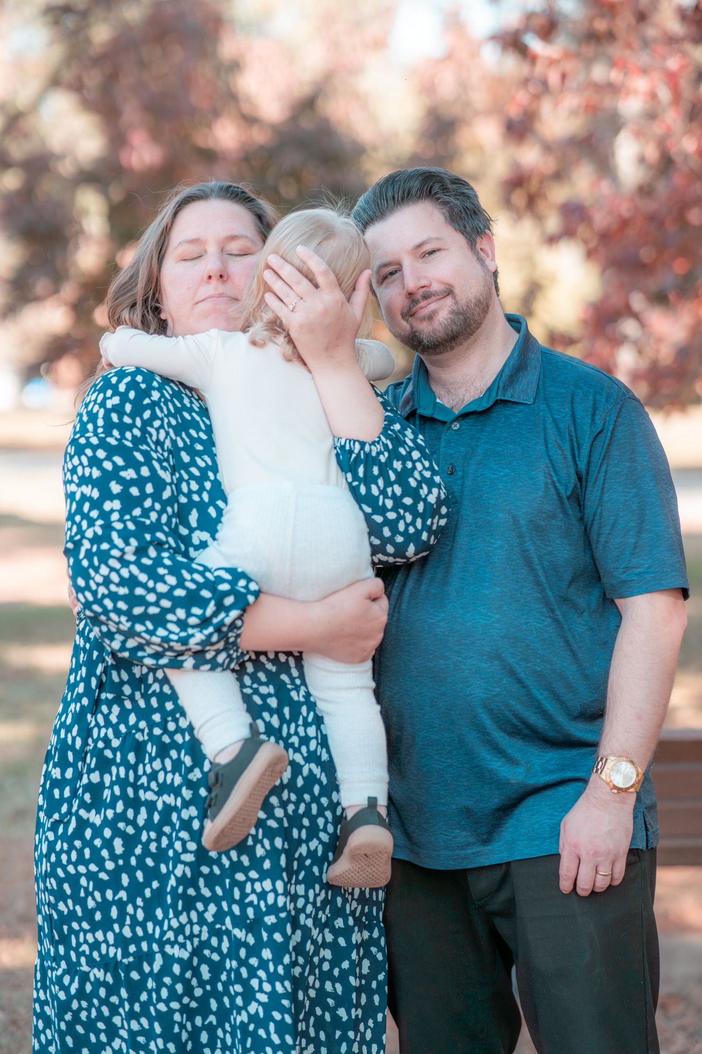 Family portrait outdoors with fall foliage background showing couple in blue attire holding small child in white.
