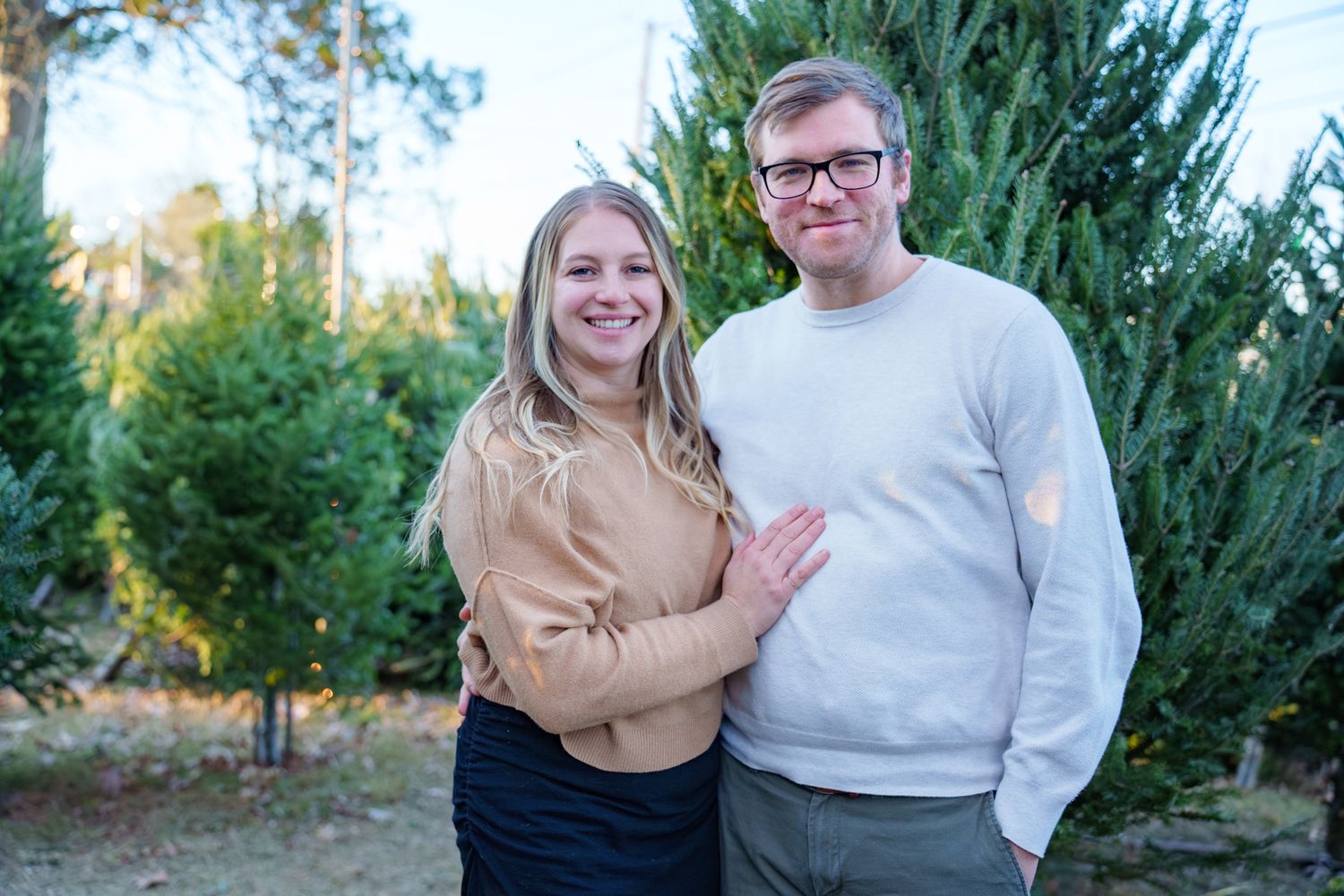 Couple poses together in front of Christmas trees at a tree farm during sunset.