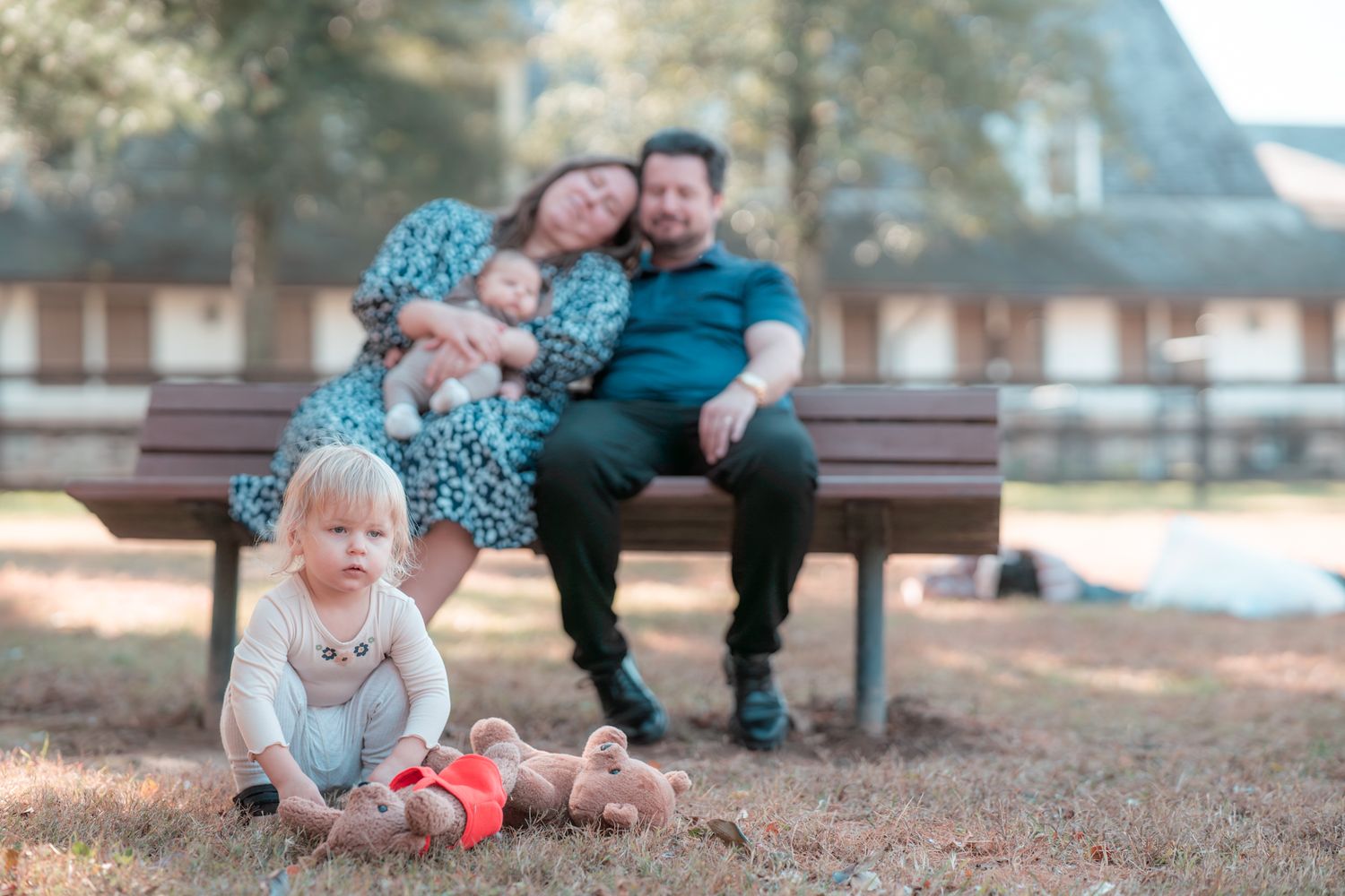 Family relaxing together on a park bench while young child plays with a ball in the foreground.