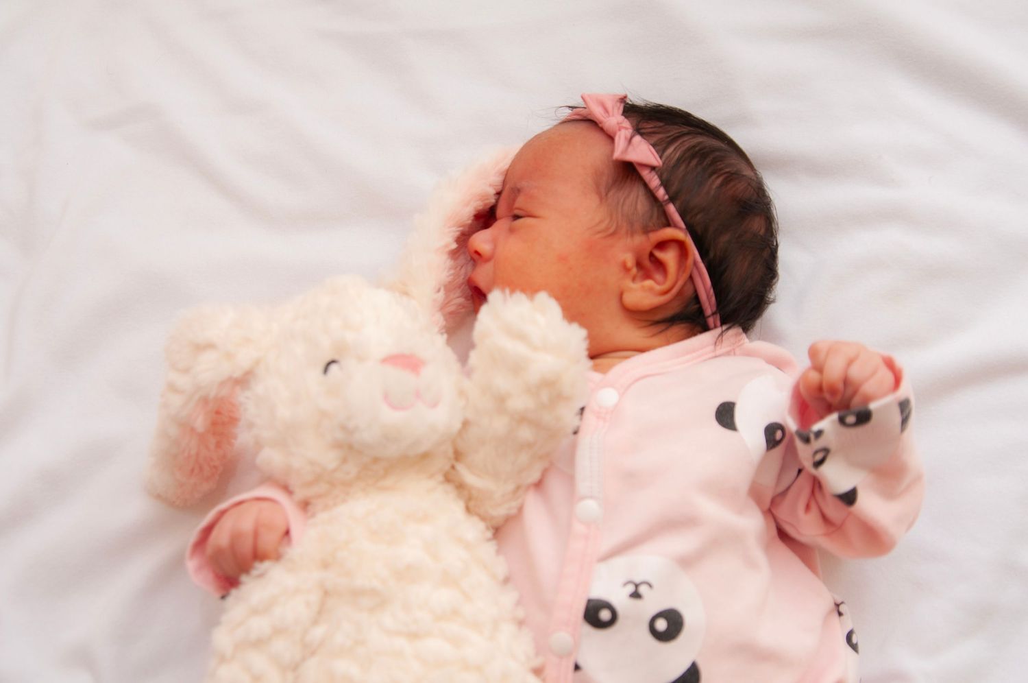 Newborn baby in pink panda outfit lying next to plush teddy bear on white bedding for a lifestyle newborn session