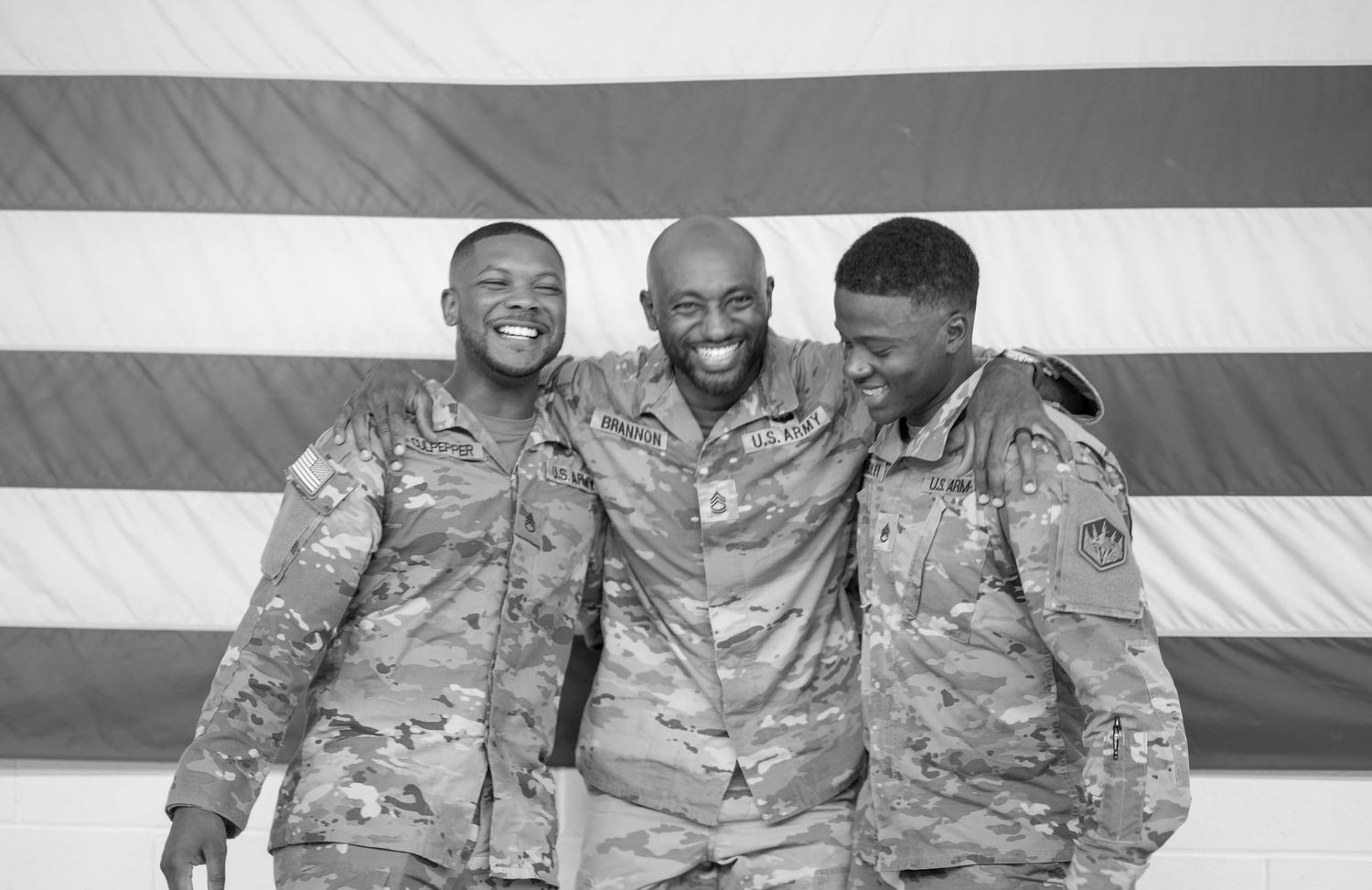 Three soldiers in military camouflage uniforms smile together in front of an American flag in black and white.