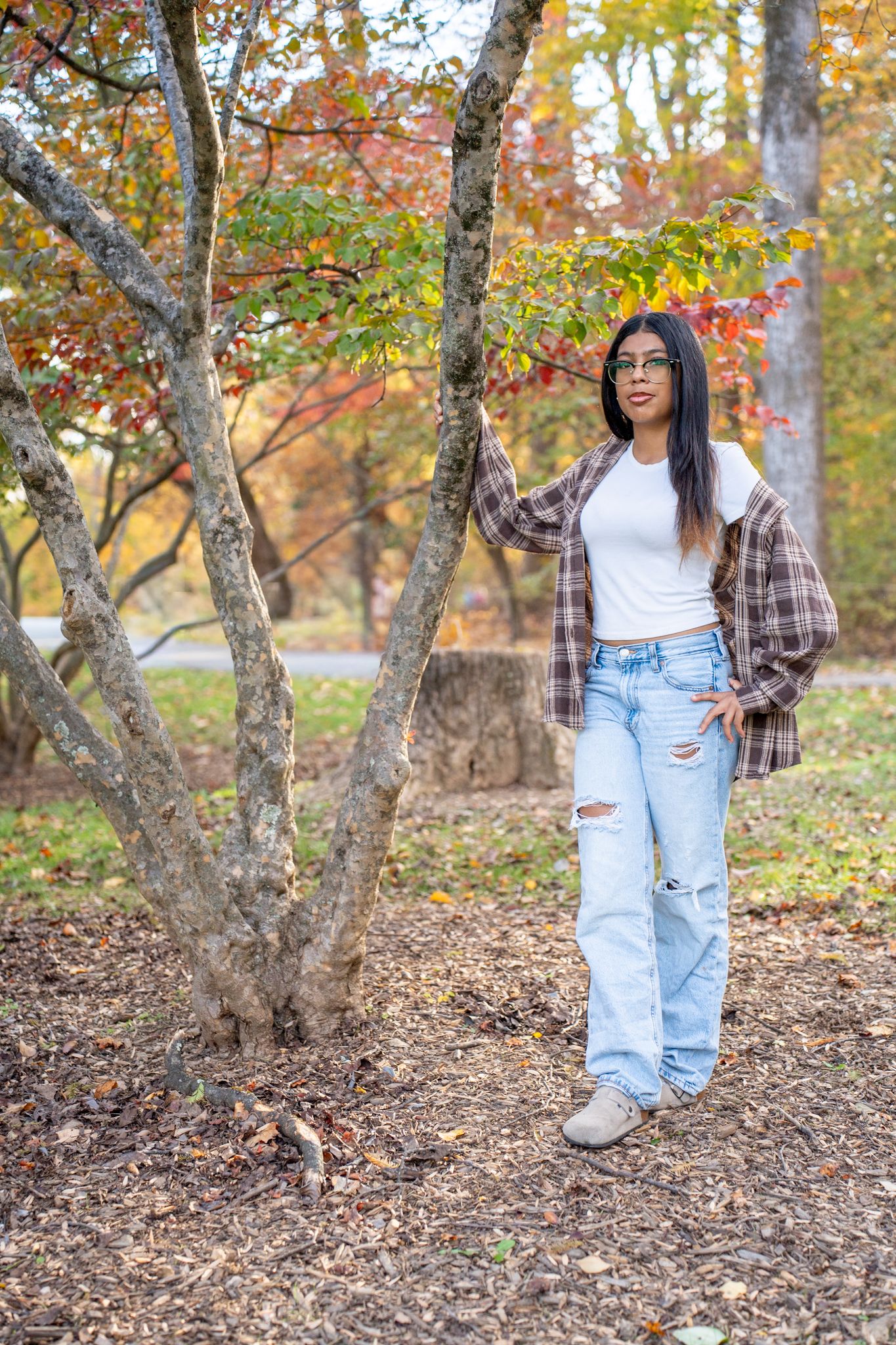 Person wearing a plaid shirt and light denim jeans poses in an autumn forest setting with colorful fall foliage.