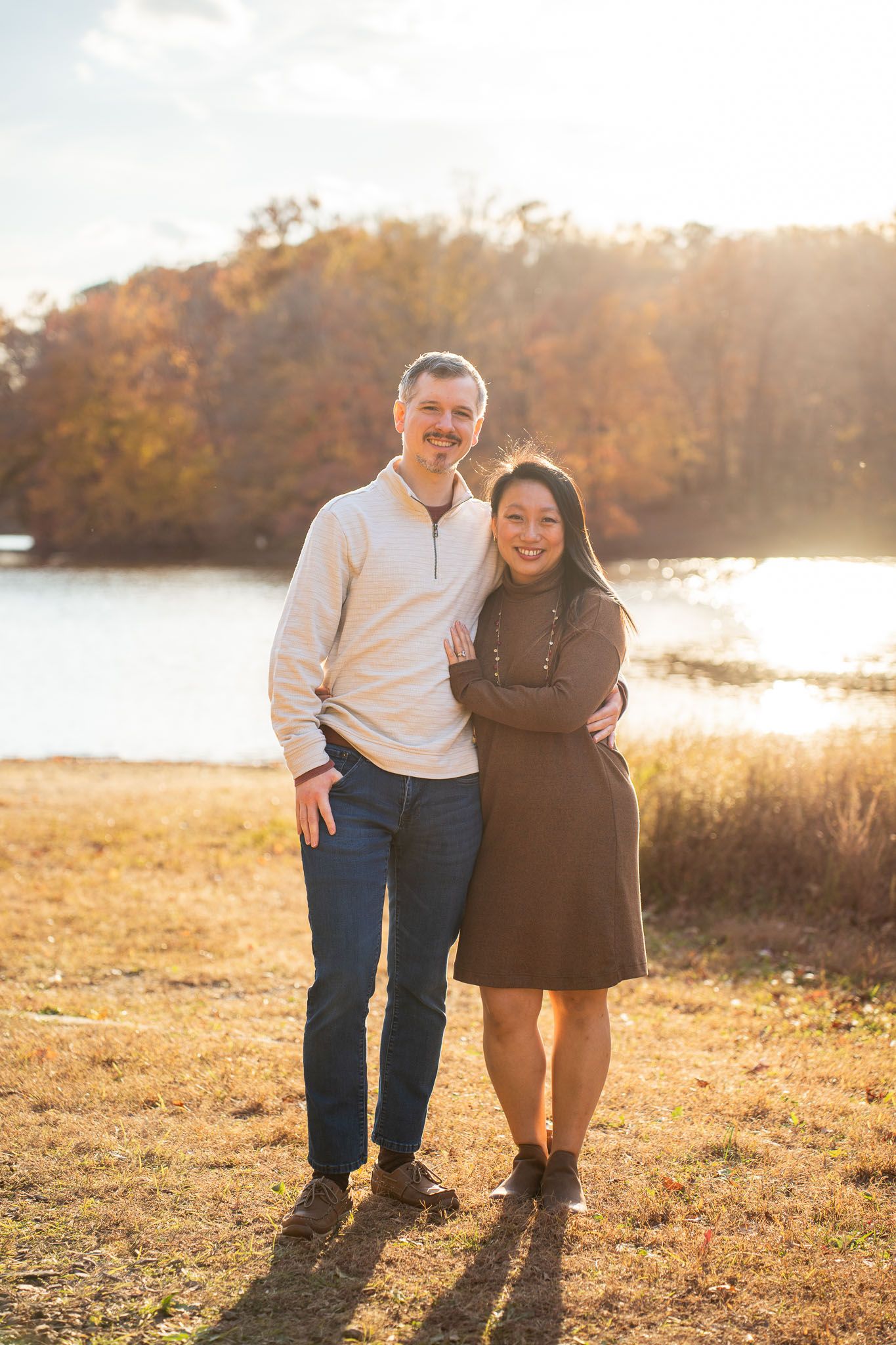 Couple embraces in warm autumn sunlight on grassy field during golden hour sunset photoshoot.