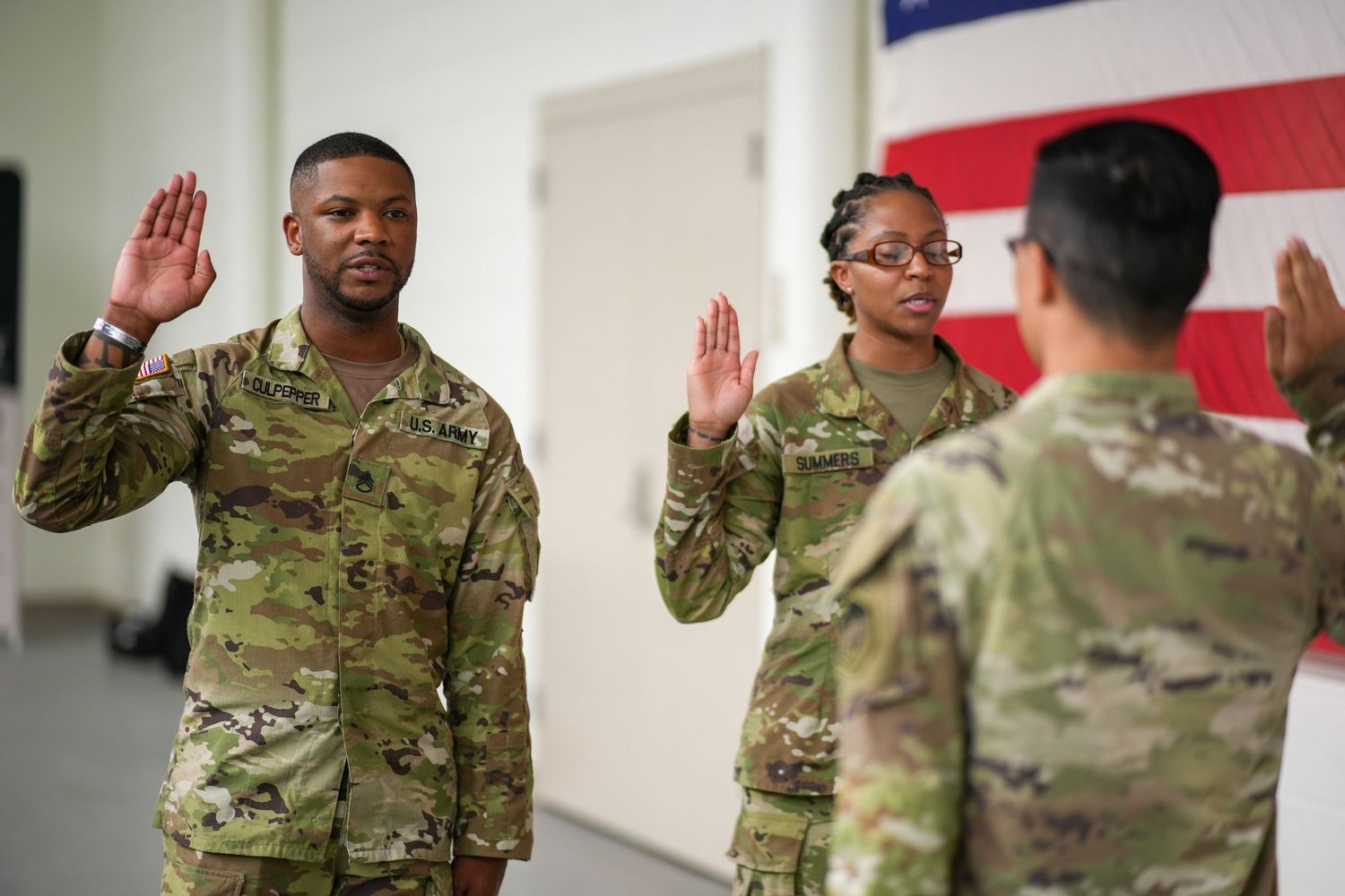 Military service members in camouflage uniforms raise their hands in an oath-taking ceremony in front of an American flag.