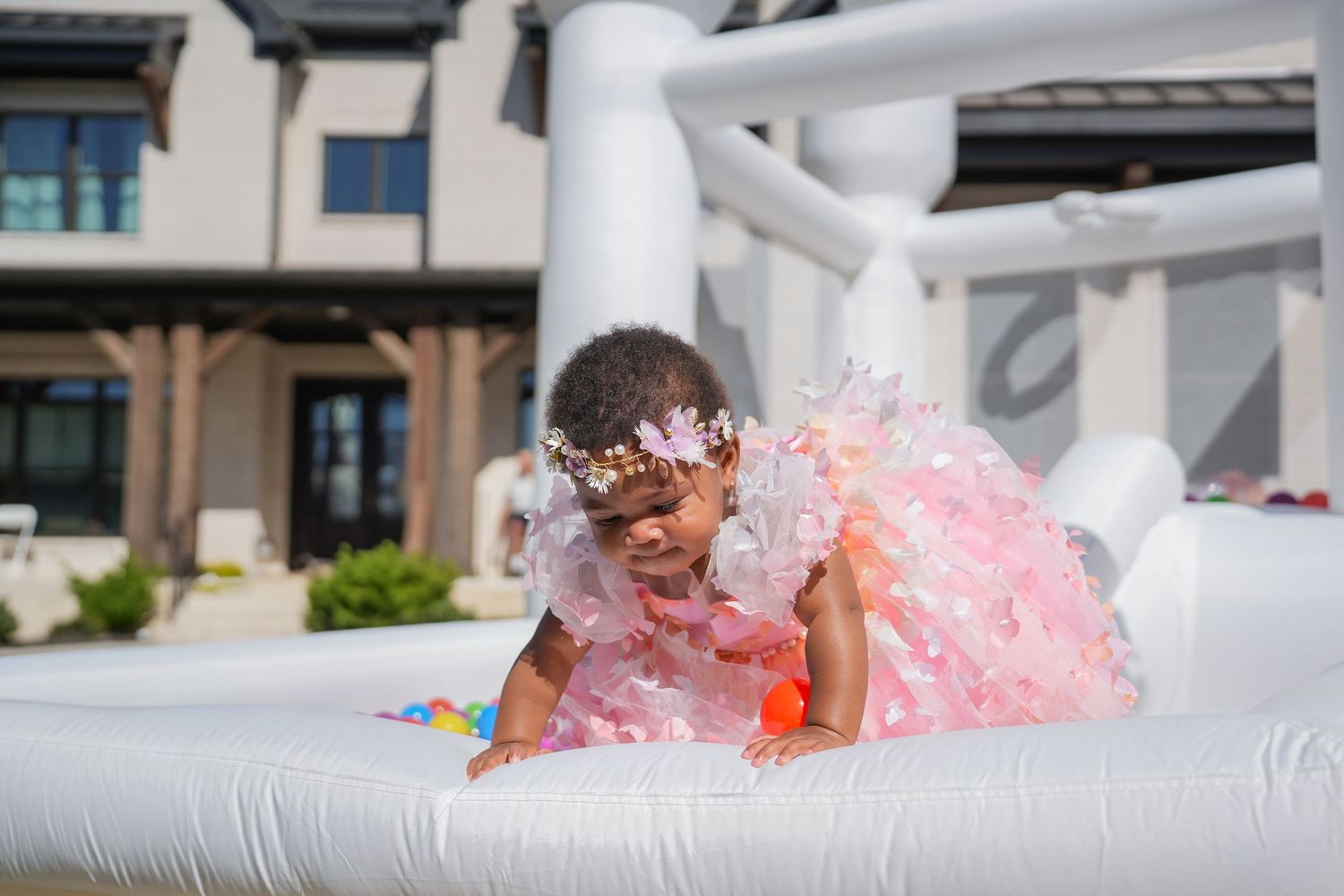 A child in a pink tulle dress crawls on a white inflatable bounce house at an outdoor party.