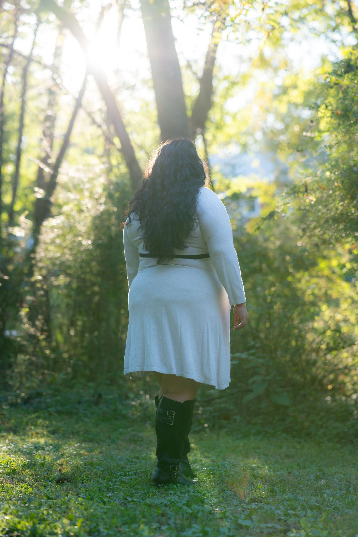 Person in a white sweater stands in a sunlit forest clearing surrounded by trees and scattered yellow flowers.
