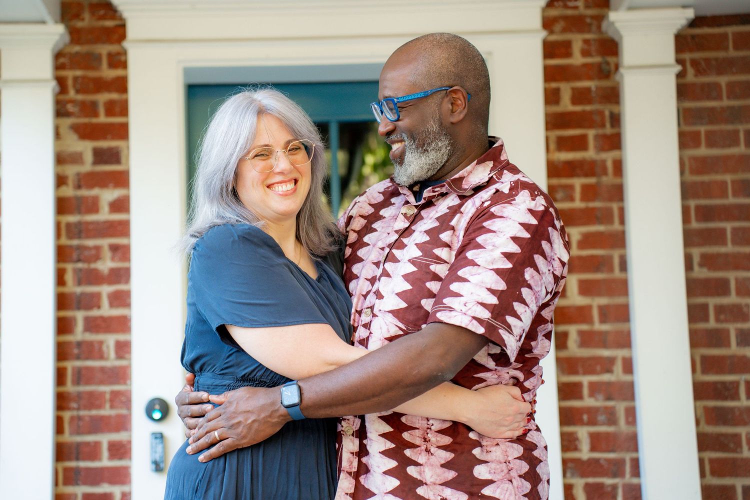 A mature couple embraces while standing on a brick porch, wearing casual attire and sharing a warm moment together.