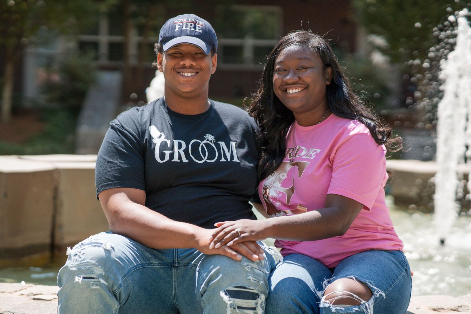A couple wearing matching groom and bride shirts sit together smiling in front of a fountain.
