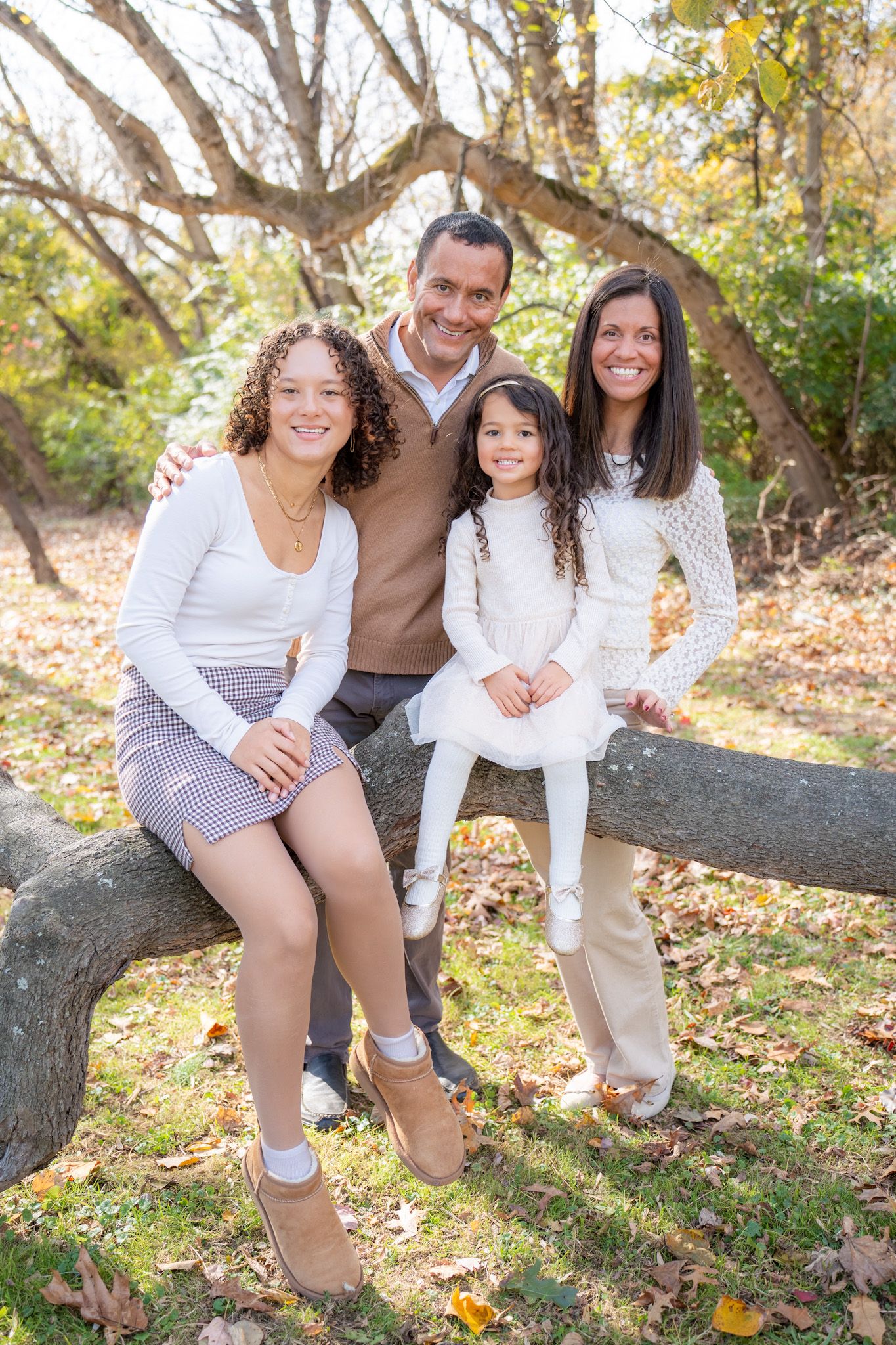 A family of four poses together on a fallen tree branch during an autumn photoshoot in a wooded setting.