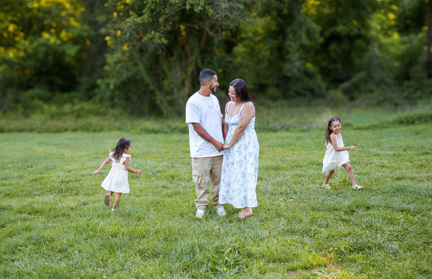 A pregnant couple holds hands in a green meadow while two young daughters in white dresses play around them.