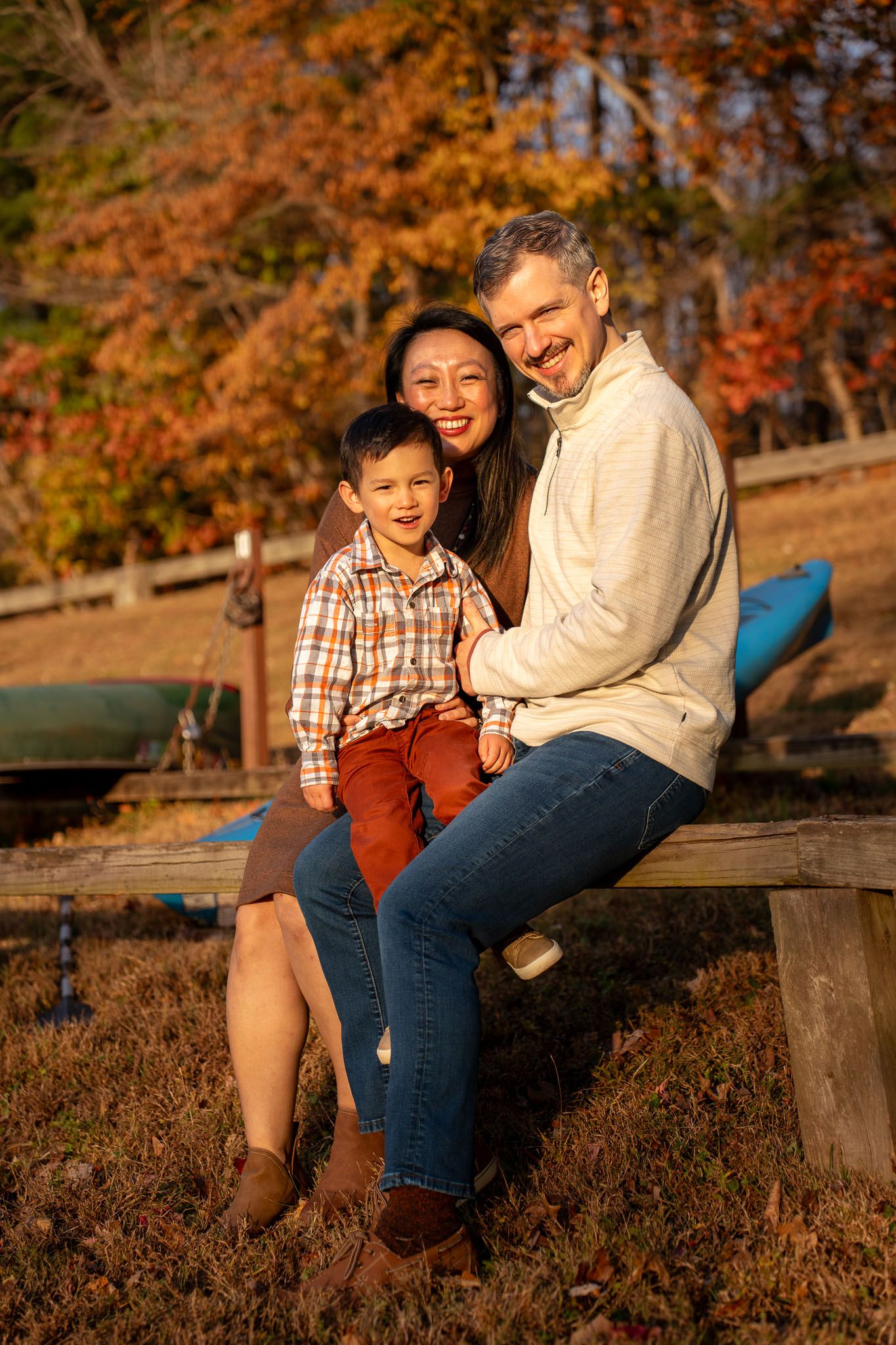 A family sits together on a wooden bench during golden hour with autumn foliage in the background.