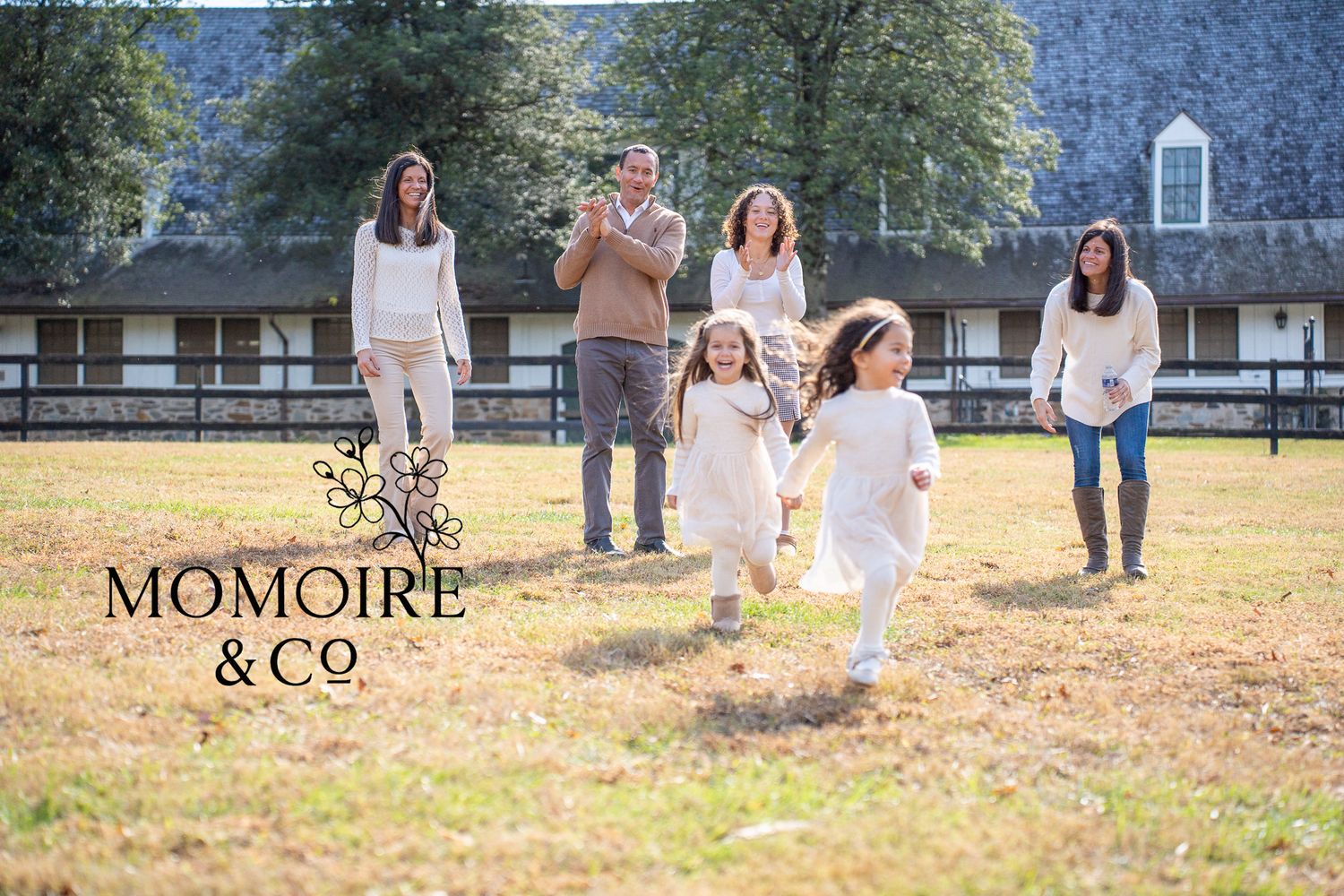 A family plays together in a sunny field outside a rustic barn with white fencing on a beautiful autumn day.