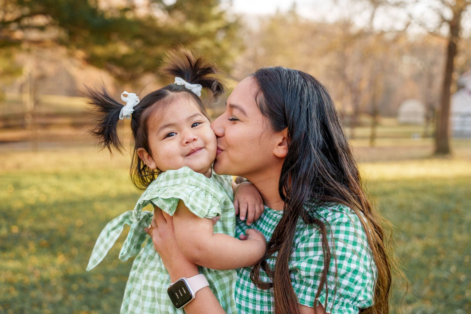 A parent and child in matching green gingham outfits share a tender moment outdoors in a sunny park.