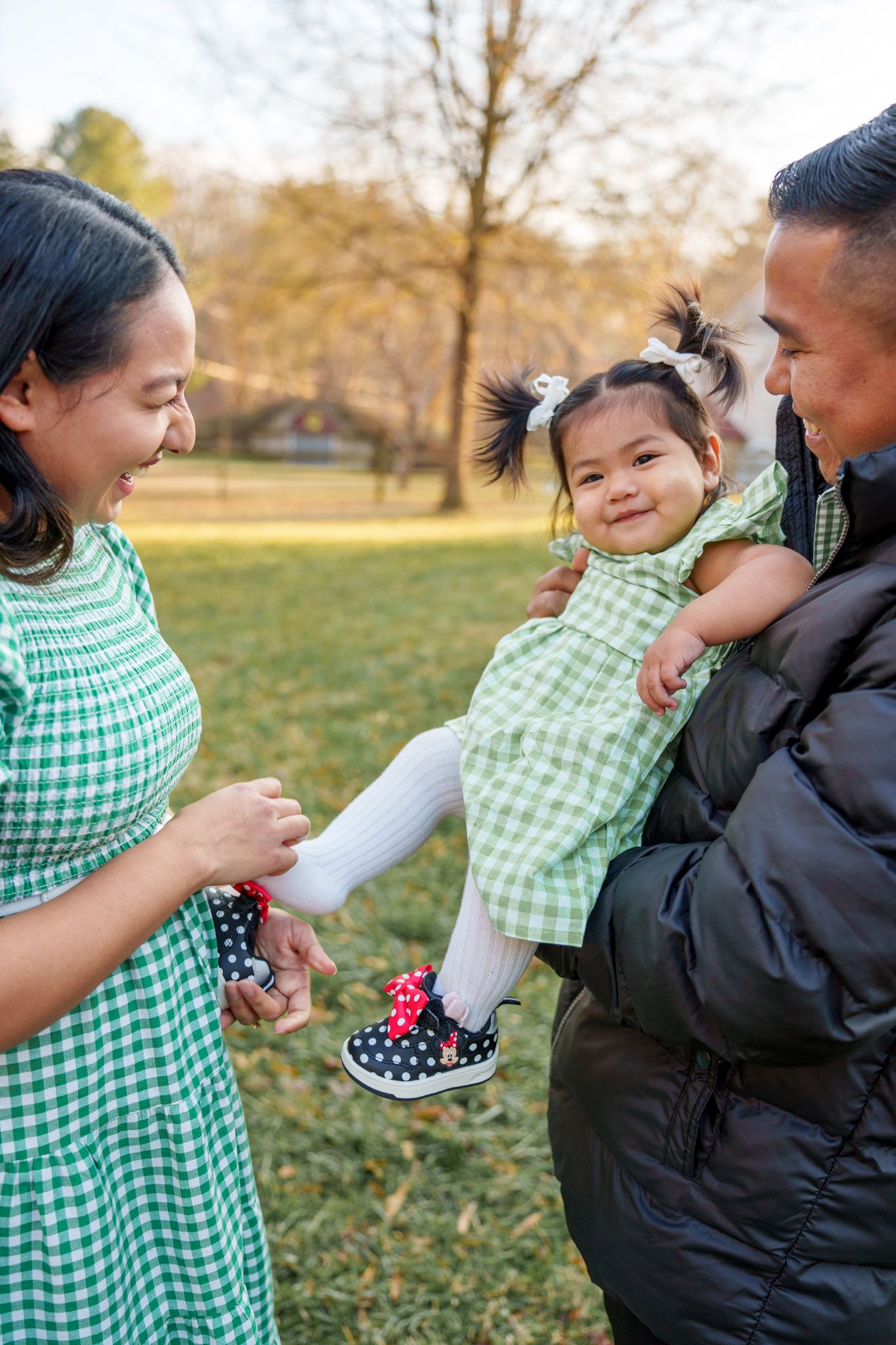 A family plays together outdoors in autumn, with a child in a green gingham dress being lifted playfully in the air.