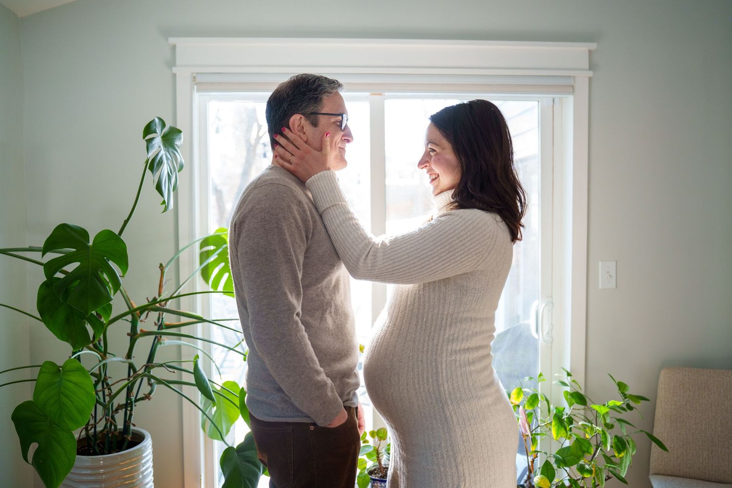 Two people stand in a bright room with houseplants near a window, sharing an intimate moment together.