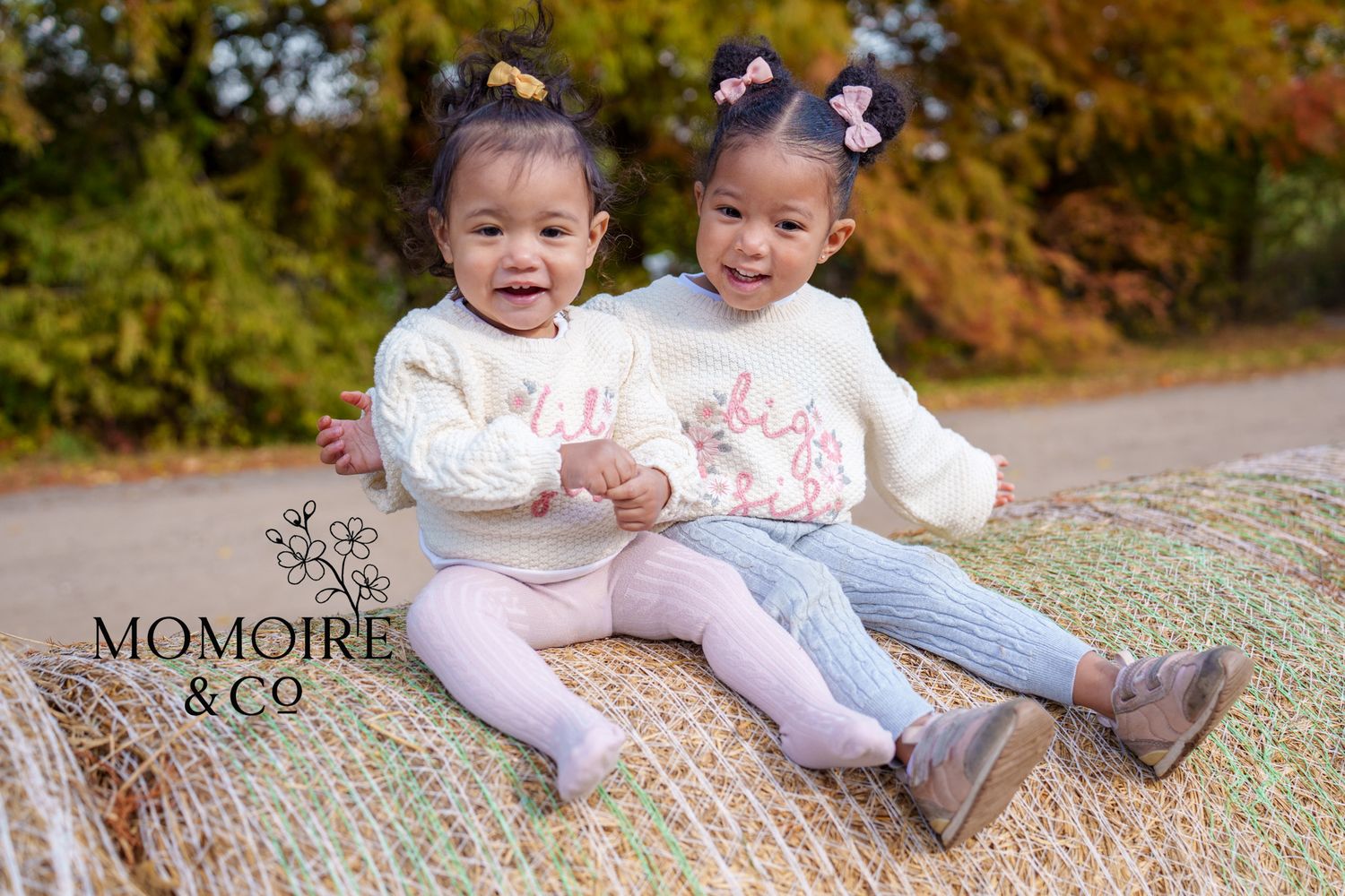 Two young children in white sweaters and leggings sit together on a blanket during an autumn outdoor photo session.