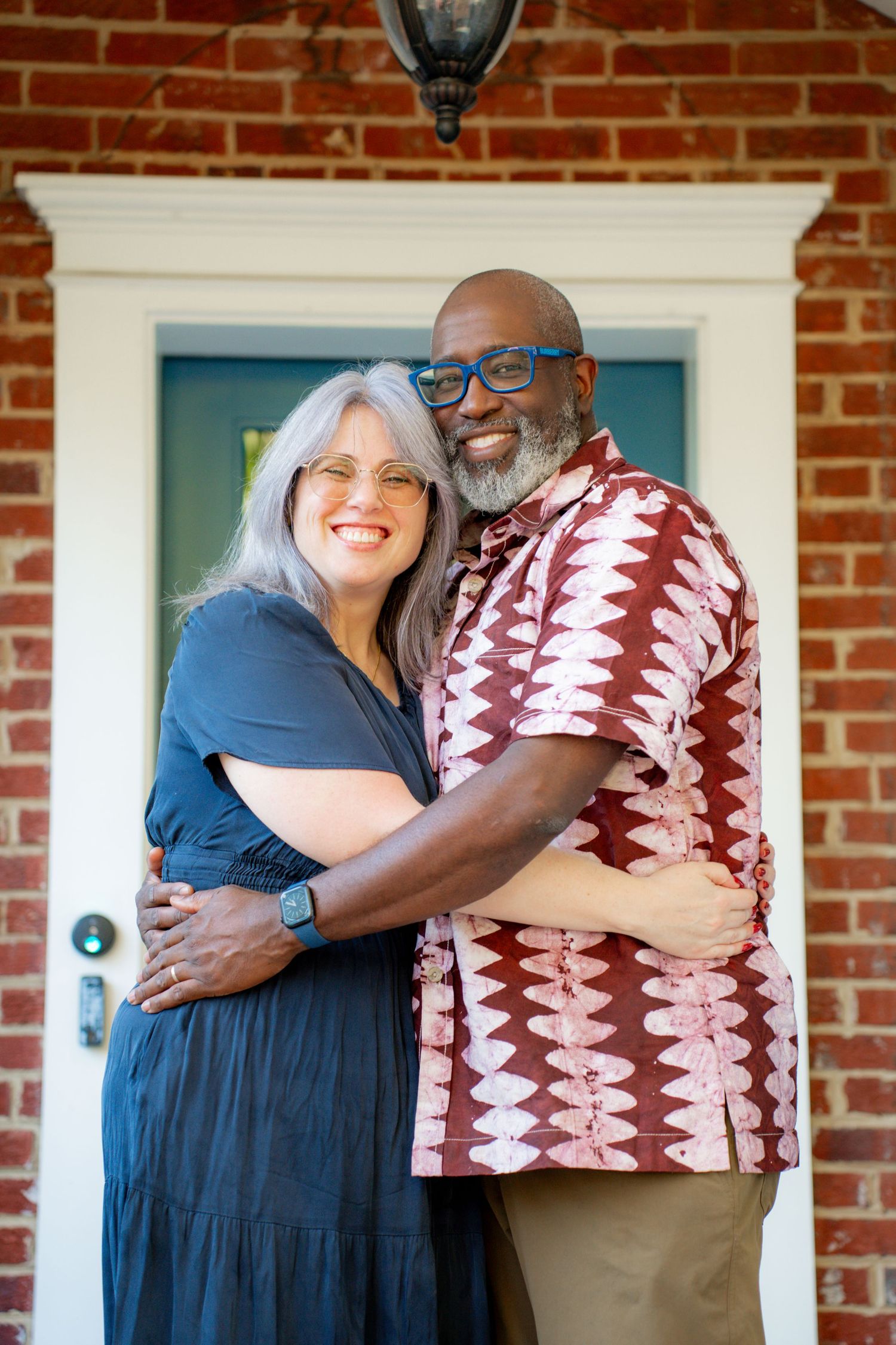 Happy couple embraces on brick porch, wearing blue dress and patterned shirt in front of white door frame.