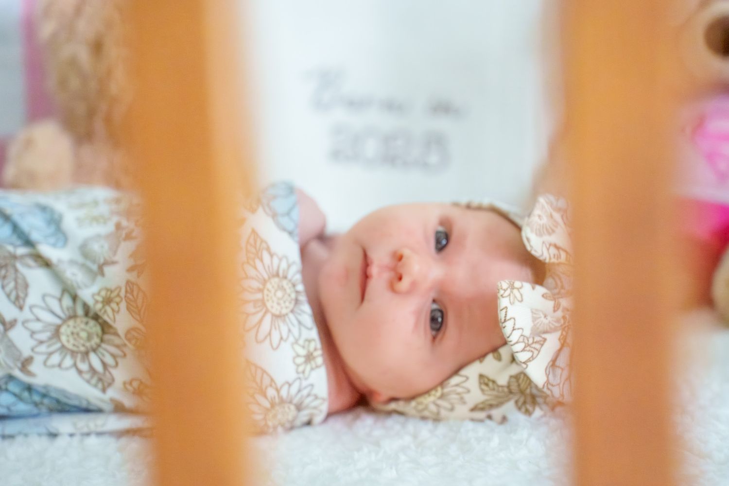 Baby lying on patterned bedding viewed through crib slats in soft lighting.