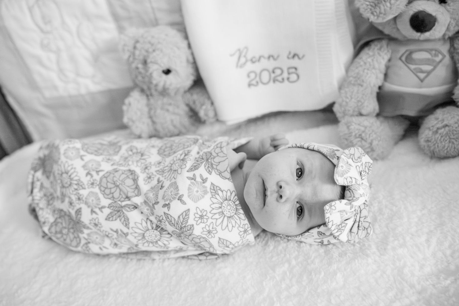 Black and white photo of a baby wearing a lace outfit lying on white bedding with stuffed animals and a Superman toy.