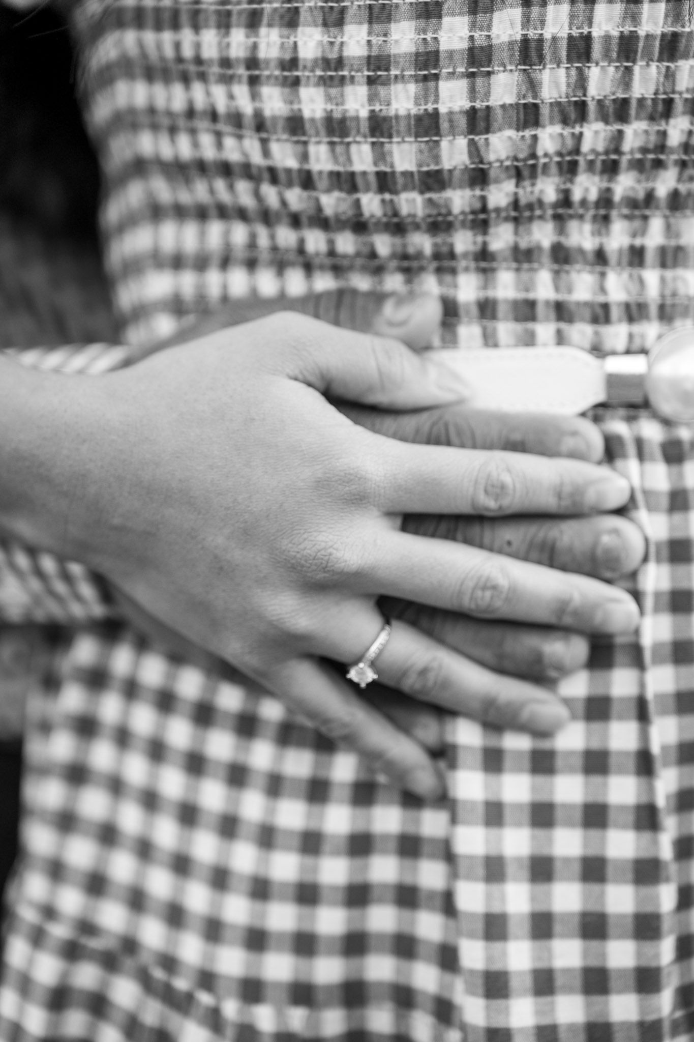 Black and white photo of couple embracing, woman's hand with engagement ring resting on partner's plaid shirt.