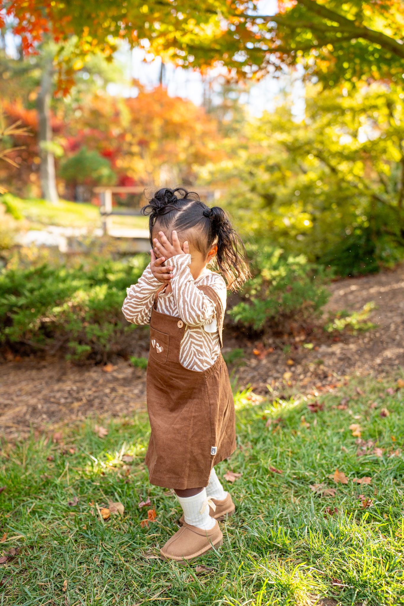 A small child plays in fallen autumn leaves wearing a brown vest and striped shirt on a sunny day.