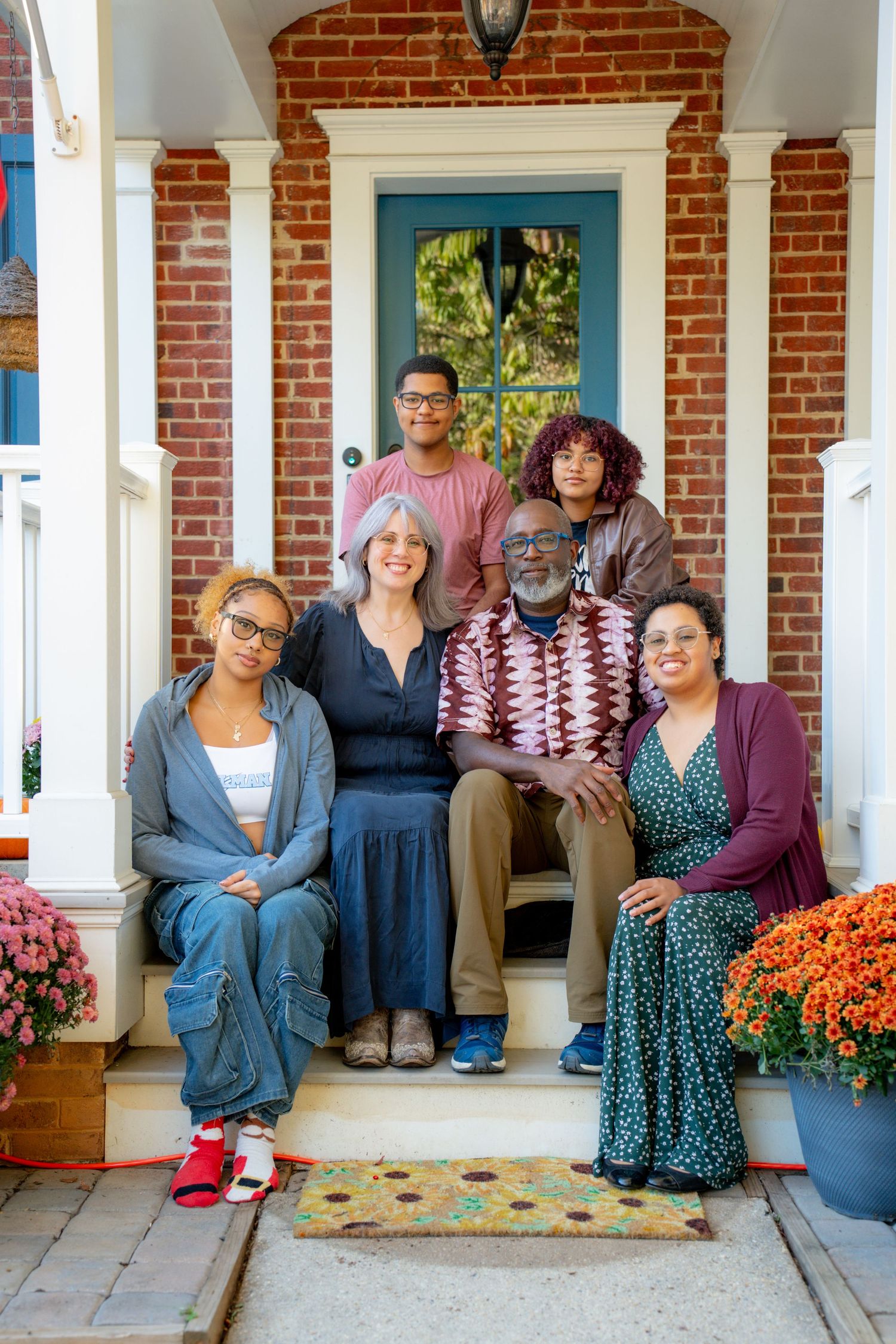 A group sits together on a porch with white columns and brick walls, decorated with orange mums and fall decor.