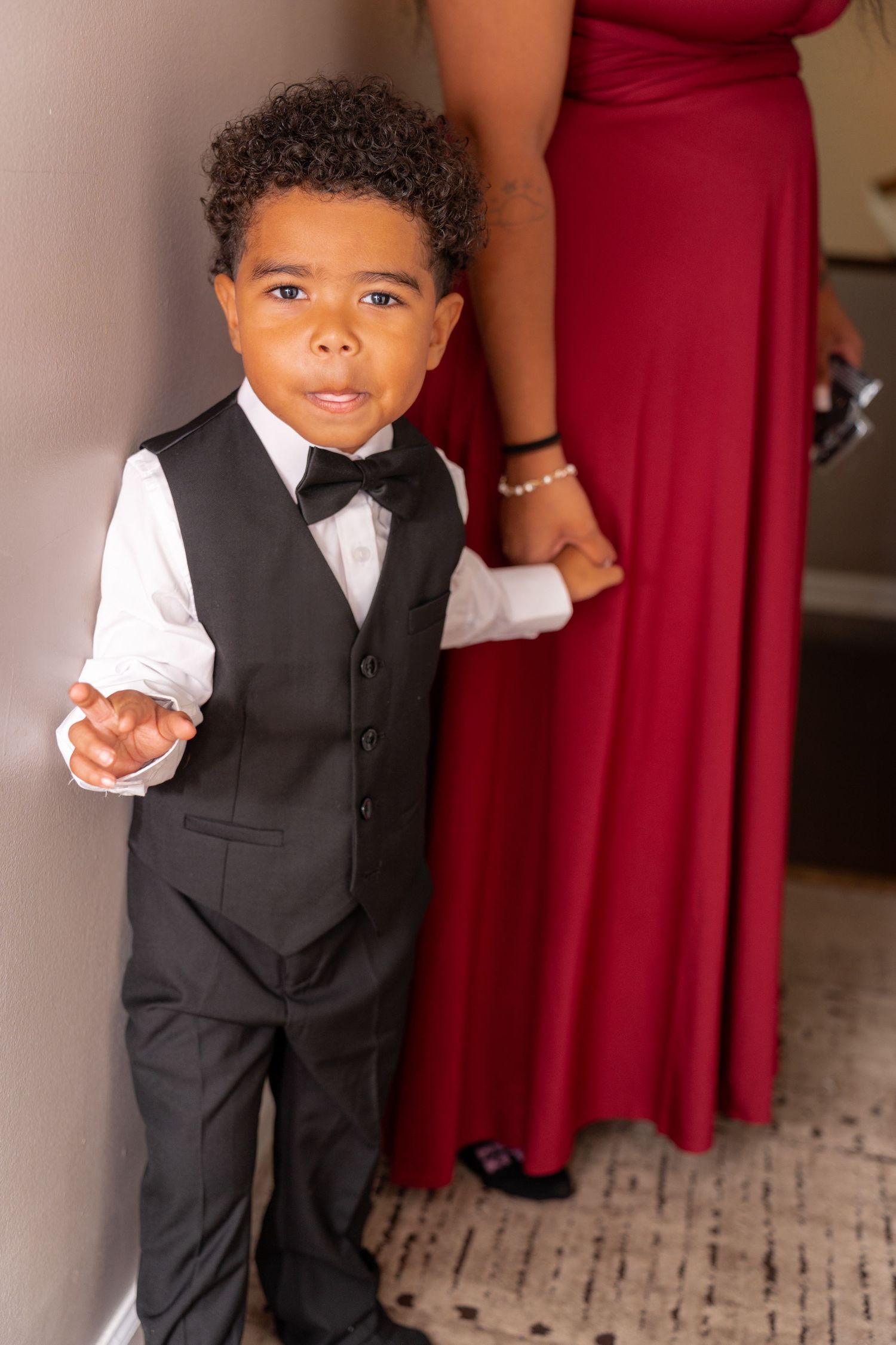 A young child in formal attire with a black vest and bowtie stands next to someone wearing a long red dress.