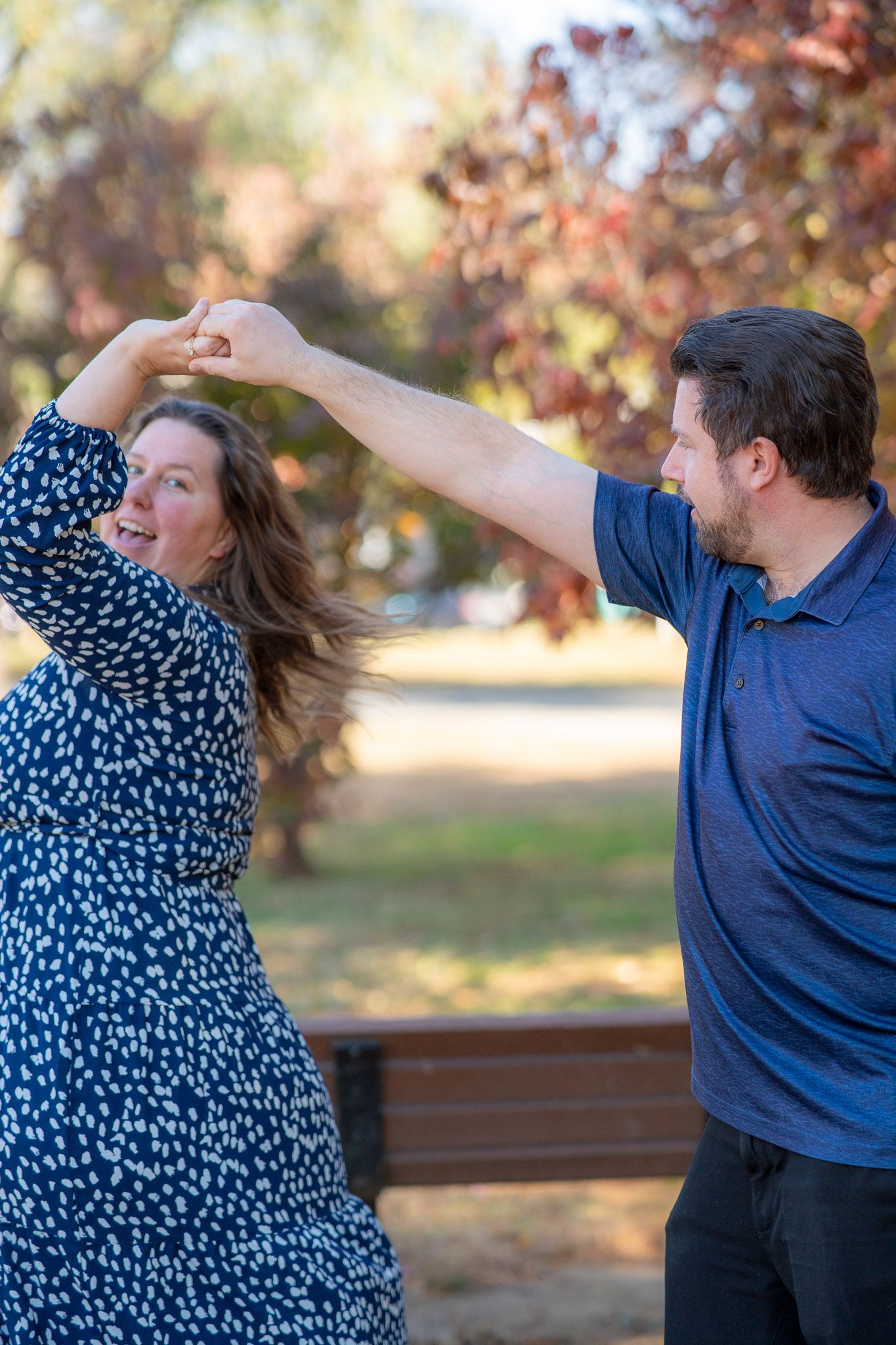A couple dances playfully outdoors in a park during autumn with fall foliage in the background.